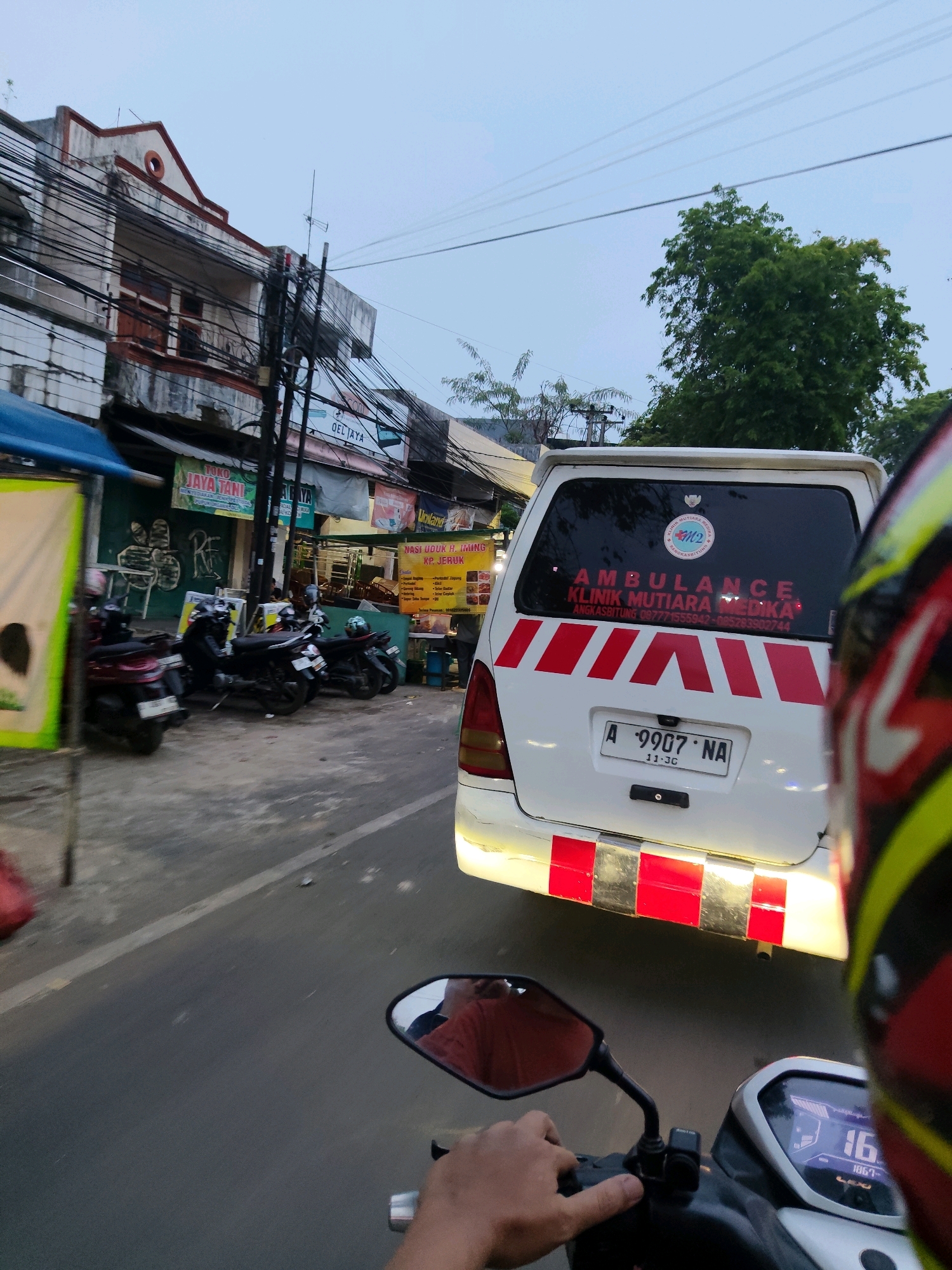 The image captures a street scene in Rangkasbitung, Indonesia, viewed from the perspective of a motorcycle rider. The foreground features the rider's hand on the handlebar and a partial view of the motorcycle's digital display, showing "16" for speed. To the right, the rider's helmet is partially visible. In the middle ground, a white ambulance with red and white reflective stripes on its rear is the most prominent subject. Text on the ambulance's rear window reads "AMBULANCE KLINIK MUTIARA MEDIKA". A license plate with "A 9907 NA" is visible. The ambulance is parked or moving slowly on a paved road.
To the left of the ambulance, several motorcycles are parked along a sidewalk. Behind the motorcycles, a row of buildings lines the street, displaying various signs. One sign says "TOKO JAYA TANI". The buildings appear to be shops or small businesses, with awnings and overhead electrical wires. Trees and utility poles are also present. The sky is overcast, suggesting it might be late afternoon or early morning. The overall impression is a typical street scene in a bustling urban environment in Indonesia.