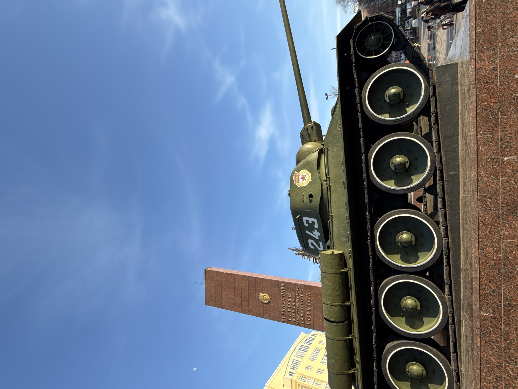 A dark green T-34-85 medium tank, marked with the white number "243" on its turret and a red star "ГВАРДИЯ" (Guard) emblem, is prominently displayed as a monument. It rests on a multi-tiered base composed of reddish-brown granite. Behind and to the left of the tank stands a tall, reddish-brown rectangular obelisk, bearing a gold-colored emblem and gold Cyrillic text that reads: "ВЕЧНАЯ СЛАВА ГЕРОЯМ ВОИНАМ ДОБРОВОЛЬЧЕСКОГО ТАНКОВОГО КОРПУСА" (Eternal Glory to the Heroes-Soldiers of the Volunteer Tank Corps). This memorial is situated at the intersection of Komsomolsky Prospekt and Oktyabrskaya Square in Perm, Perm Krai, Russia. A multi-story building with a light yellow facade is partially visible in the left background. The sky is clear and light blue, with minimal cirrus clouds and a visible crescent moon in the upper left quadrant. Several indistinct individuals are present in the far right background, at ground level. The scene is illuminated by daylight.