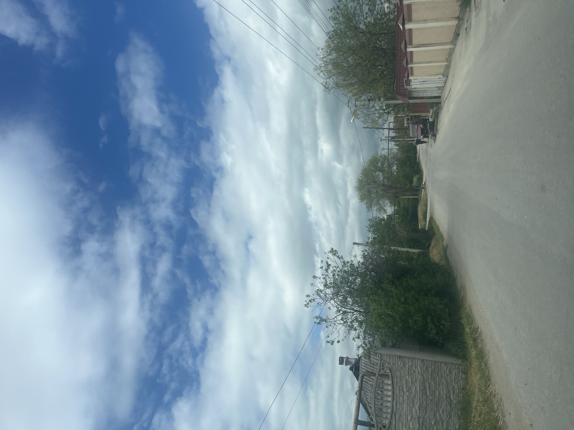 The image depicts a street scene in an unknown urban location. The view is down a gray paved road, bordered on one side by buildings and on the other by vegetation and a fence. The sky is visible overhead, showcasing a mix of blue patches and cloud cover. Power lines run above the street.