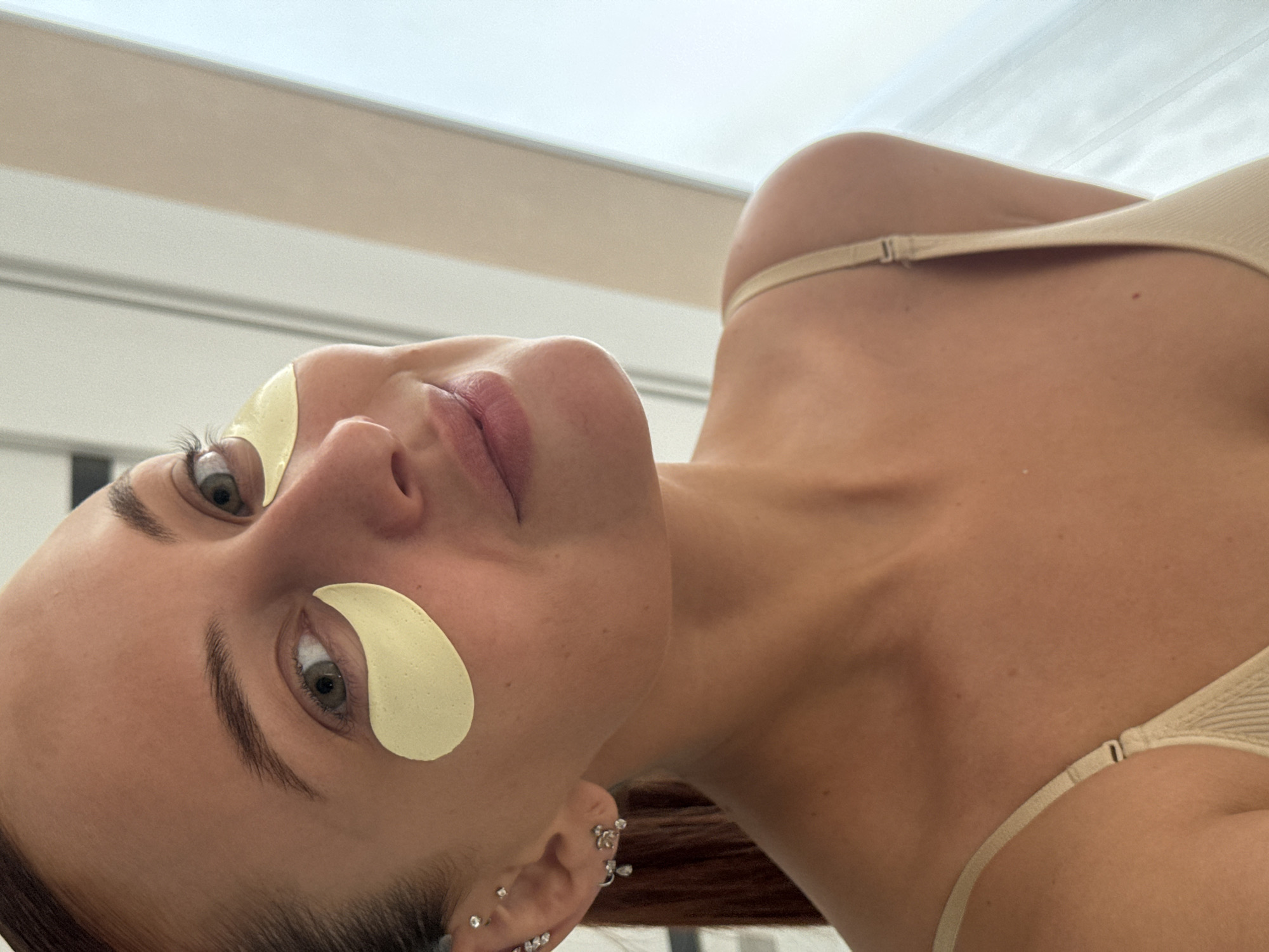 A woman is lying down, with eye patches on. She has fair skin, light eyes and is wearing a top with spaghetti straps. The background appears to be an indoor setting. The image was taken with a low angle, looking up at the subject. There is no clear evidence to confirm the location of the photo as Chaikovsky, Russia.