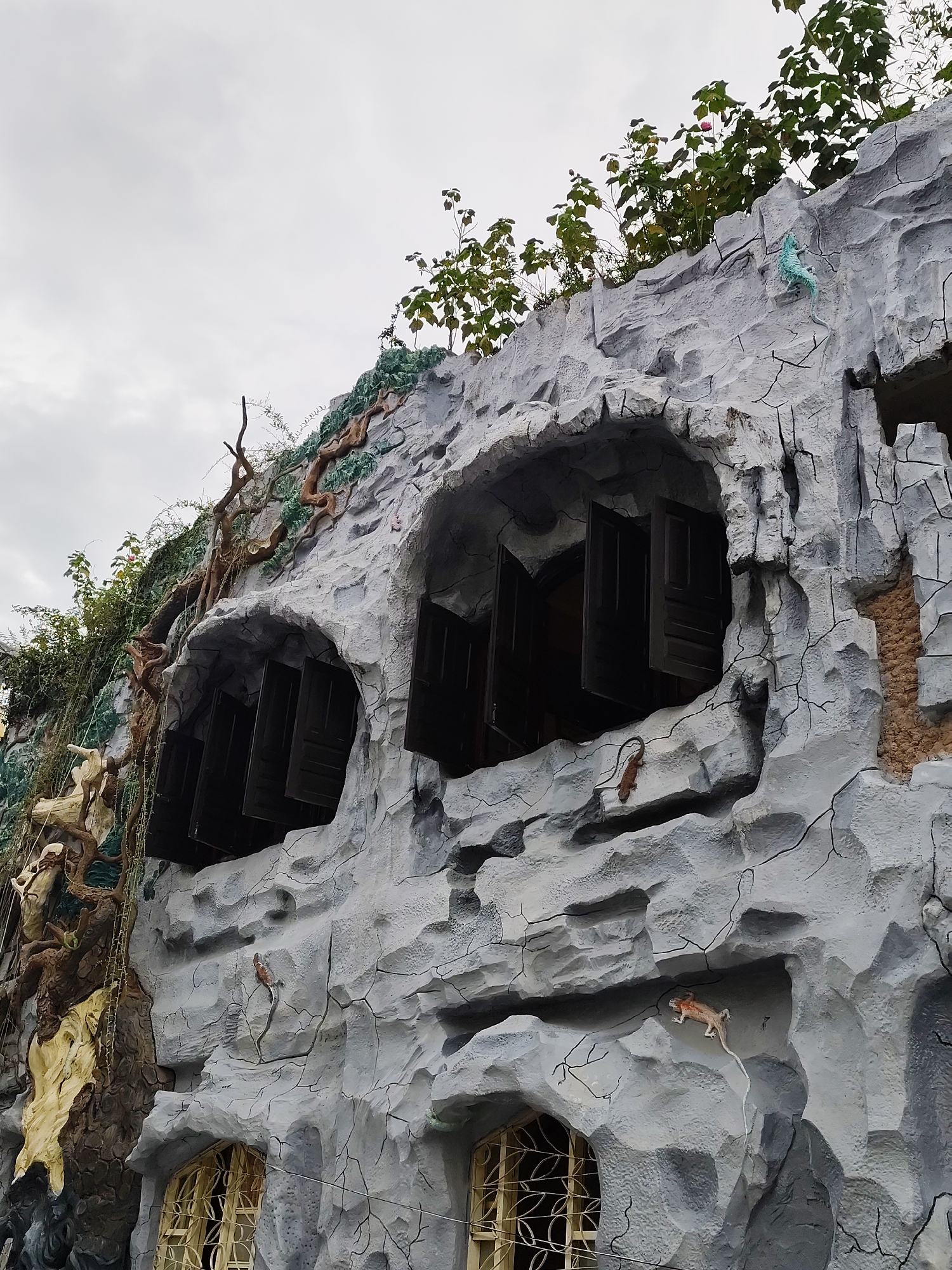 The image shows the exterior of a building designed to resemble a cliff face with stylized rock formations. Several windows with dark shutters are visible, recessed into the faux rock. Two small, decorative lizards are placed on the rock surface, one light brown and the other light blue. Vegetation, including vines and leafy branches, grows around and on top of the structure. The sky is overcast and gray. This building is located in Da Lat, Vietnam, and is known as the Crazy House or Hang Nga Guesthouse.
