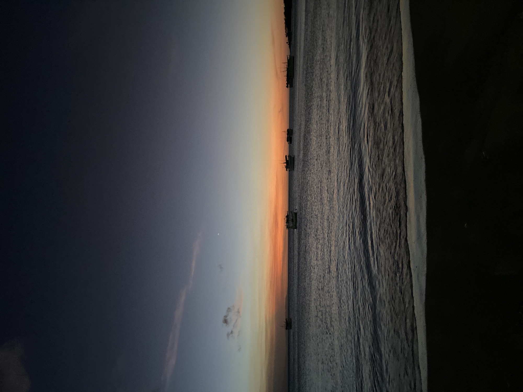 This image depicts a coastal scene at dusk in Juan Griego, Venezuela. The foreground consists of a dark, wet sandy beach, meeting the ocean with a thin line of white foam from breaking waves. The mid-ground features a body of dark, undulating water, consistent with ocean waves. Approximately five boats are visible across the water, appearing as dark silhouettes against the horizon. These vessels exhibit varying superstructures, suggesting they are fishing or small transport boats. The horizon is marked by a vibrant band of orange, red, and yellow light, indicative of a sunset or sunrise. Above this band, the sky transitions from a lighter blue-purple directly above the horizon to a deep, dark blue overhead. A faint crescent moon is discernible in the upper left quadrant of the sky, with sparse, wispy clouds present. A distant, dark landmass is visible on the horizon to the right, silhouetted against the sky. No individuals or specific actions are discernible.
