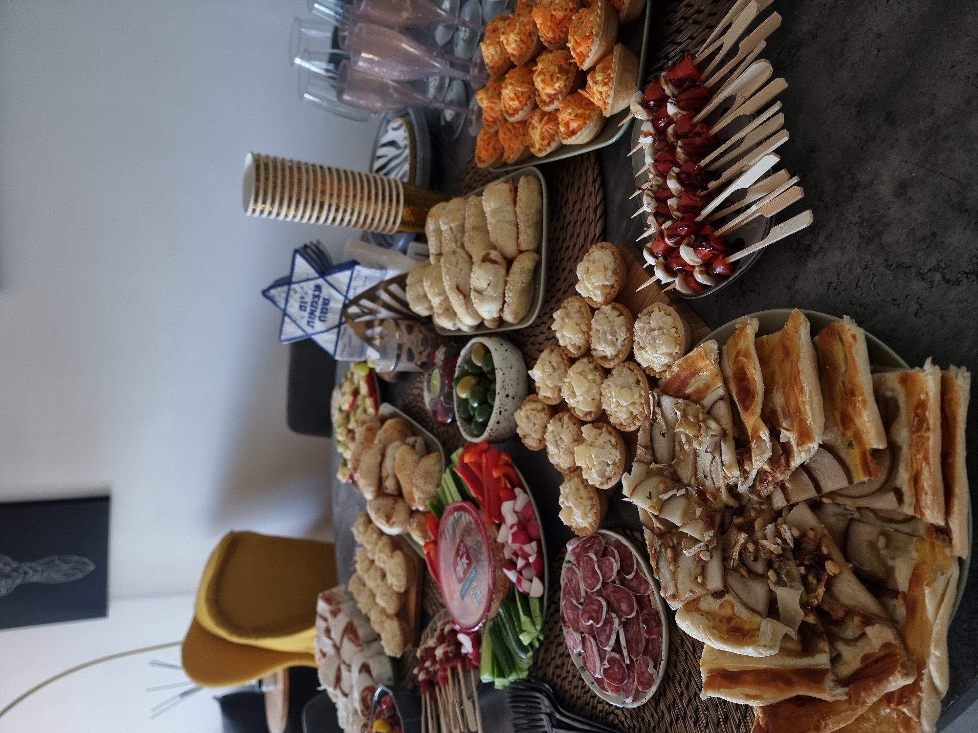 This image captures an overhead view of a festive table laden with a diverse spread of appetizers and snacks, likely prepared for a celebration or gathering. The setting appears to be indoors, possibly a dining area, with natural or bright artificial lighting.
A wide variety of finger foods are presented on different platters and dishes. These include numerous mini pita bread pockets filled with an orange, shredded mixture (possibly carrot salad), and skewers featuring red bell peppers, mushrooms or cheese, drizzled with a reddish sauce. There are also small, round pastry tarts topped with a creamy white filling, and large rectangular slices of flatbread or focaccia, some garnished with pine nuts and thin slices of what appear to be caramelized onions or fruit.
Further items include various types of small buns or bread rolls, some sprinkled with sesame seeds, and a platter of fresh, sliced vegetables (red bell peppers, cucumbers, radishes) accompanied by a red dip in a plastic container. A bowl of mixed green and black olives, a plate of thinly sliced cured meats (likely salami or sausage), and another dish containing a mixed salad are also visible.
For beverages, there's a stack of gold-rimmed paper cups and several pink-tinted champagne flutes. The table is adorned with woven placemats or mats. A notable decoration is a blue and white Star of David, a Jewish symbol, which prominently displays the Hebrew words "MAZAL TOV" (meaning "Congratulations" or "Good Luck"). This detail, combined with the elaborate food spread, strongly suggests a Jewish celebration or event, such as a Bar/Bat Mitzvah or a holiday gathering.
In the background, a dark framed artwork hangs on a light-colored wall, and part of a yellow chair is visible. No people are present in the frame.