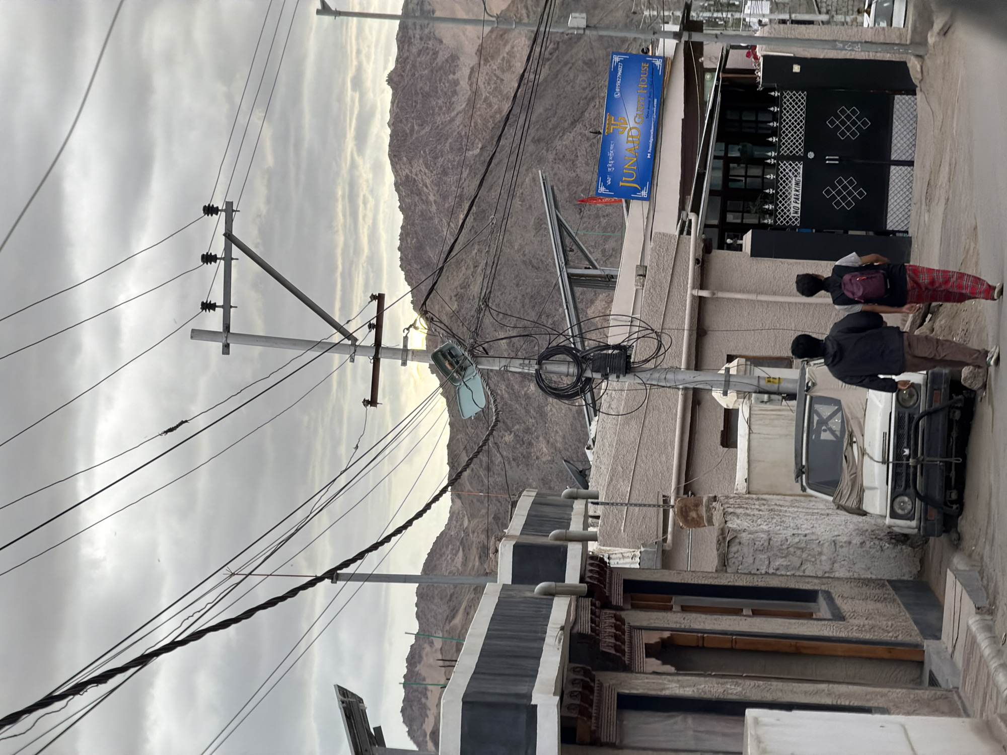 The image depicts a street scene in Leh, India, under an overcast sky. The foreground features a narrow street with a white, rugged vehicle parked on the left, covered with a tan tarp. Adjacent to the vehicle is a building with a textured facade and decorative wooden window frames.
Further into the scene, a prominent utility pole stands, entangled with a complex network of electrical wires. A blue sign for "JUNAID GUEST HOUSE" is visible, mounted on a building with a dark gate displaying a "NO PARKING" sign. Two individuals, a man and a woman, are walking away from the camera on the street, towards the guest house. The background is dominated by a rugged, mountainous landscape, characteristic of Leh. The overall atmosphere is subdued, reflecting the cloudy weather.