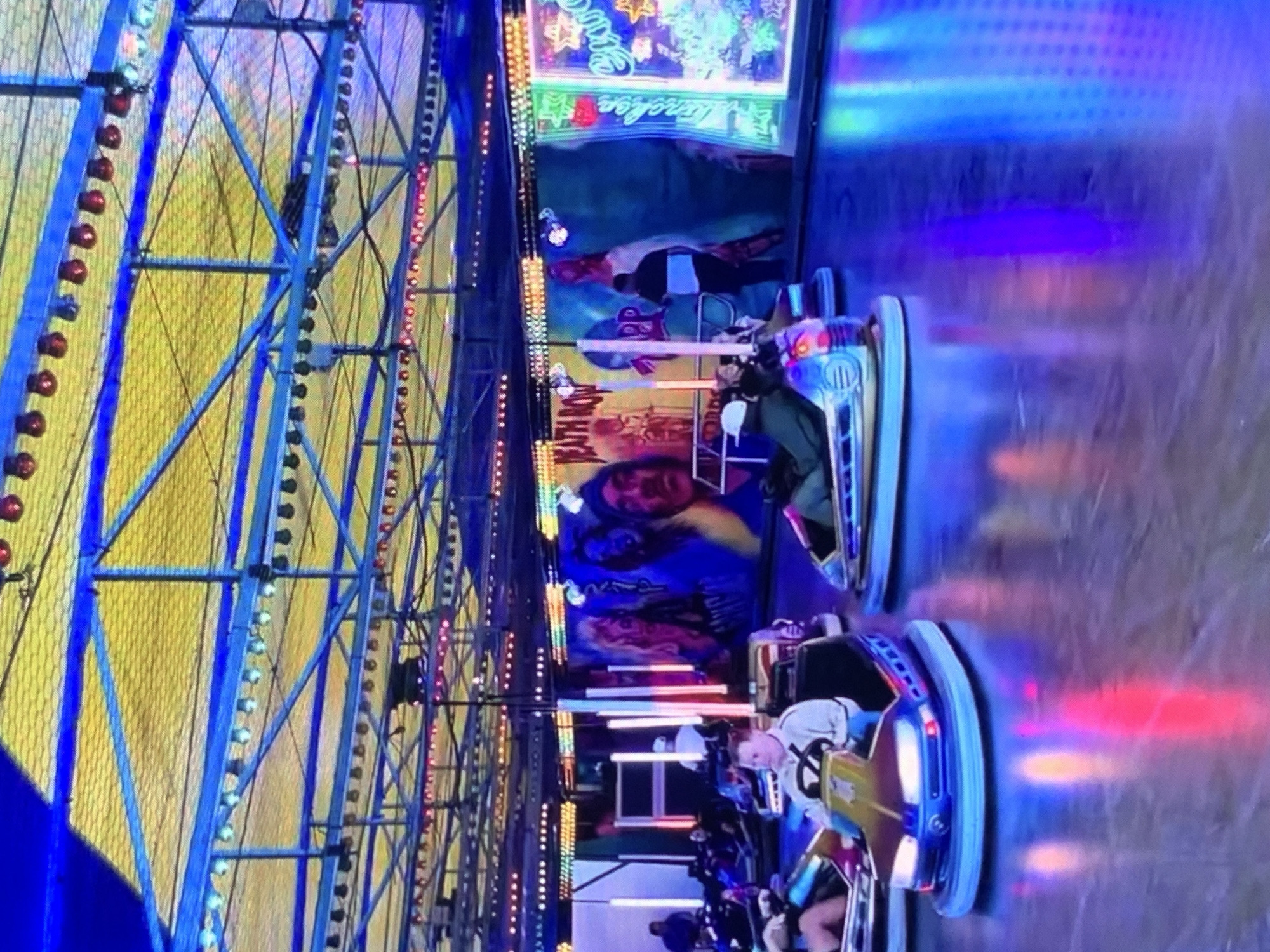 The image depicts a bumper car arena at an amusement park, likely part of a fair or carnival. The overhead structure is a complex network of blue metal beams supporting a yellow, translucent netting, with rows of colorful lights illuminating the space. Several bumper cars are visible on the polished floor, some occupied by individuals driving them. The cars are modern, with headlights and sleek designs. The walls of the arena are adorned with vibrant, illuminated signage and murals, including one that appears to read "DEATH ROW" and another with neon lights. The floor reflects the colorful lights from the cars and the surrounding decorations, creating a dynamic and energetic atmosphere. The overall scene suggests a lively entertainment venue, filled with the visual stimulation of lights and the activity of the bumper cars. There is no specific information in the image to indicate the location is Stavropol, Russia.