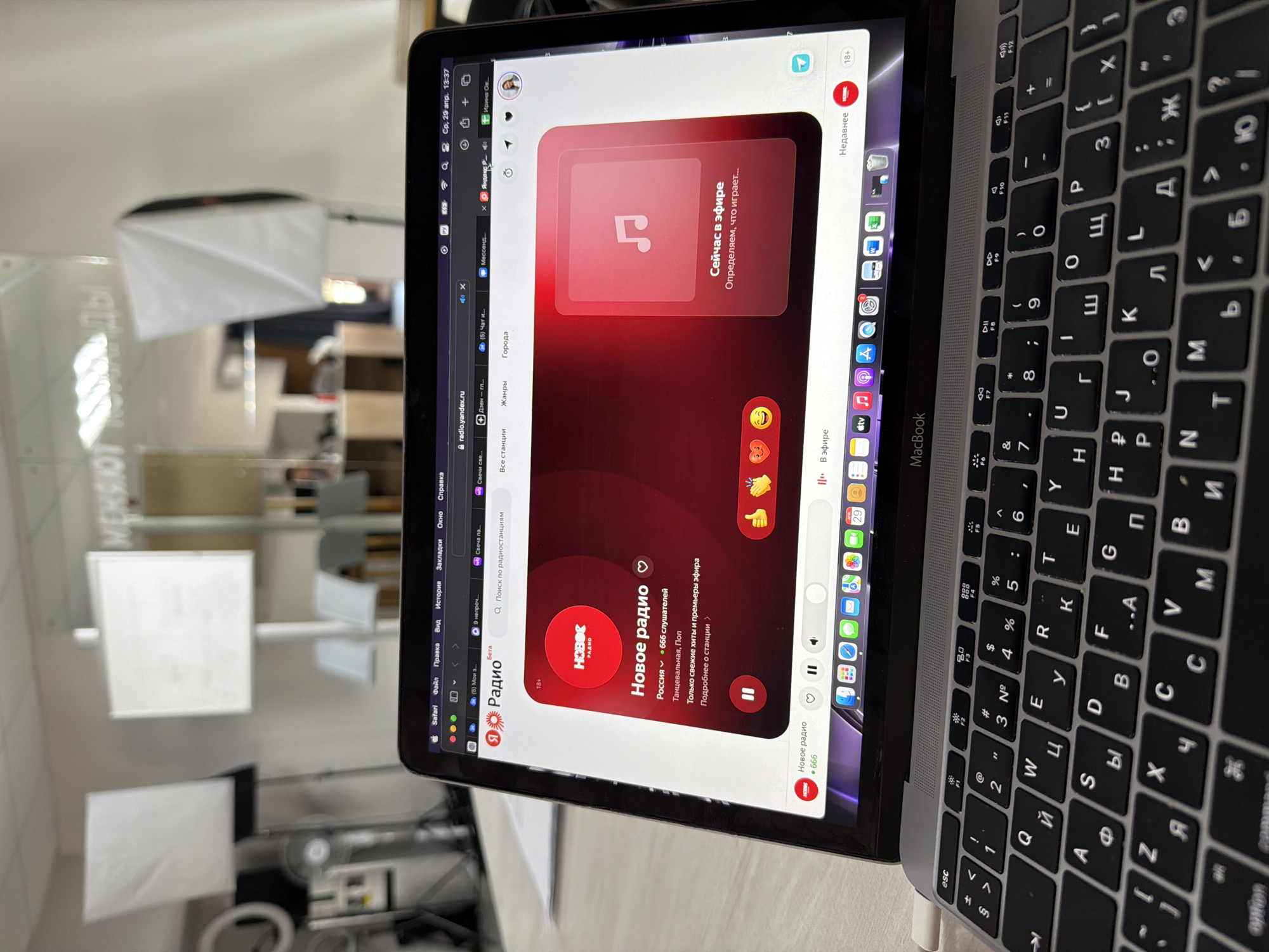 An angled shot captures a MacBook laptop on a light-colored surface, revealing its screen and a portion of the Cyrillic keyboard. The laptop screen displays the Safari web browser, currently open to "radio.yandex.ru," a Russian online radio platform. The main content area shows "Новое радио" (New Radio), indicating "666 слушателей" (666 listeners) from "Россия" (Russia), with genres "Танцевальная, Поп" (Dance, Pop). Text "Только свежие хиты и премьеры эфира" (Only fresh hits and premieres on air) is also visible. A prominent red box on the screen says "Сейчас в эфире" (Now on air) and "Определяем, что играет..." (Determining what is playing...). Emojis like a heart, fire, clapping hands, and thumbs up are displayed below the main radio content.
Other elements on the screen include the date and time "Ср, 29 апр. 13:37" (Wednesday, April 29, 13:37), a user profile picture labeled "Ирина Ов" (Irina Ov), and multiple browser tabs and macOS Dock icons for various applications. The keyboard has both Latin and Cyrillic characters.
In the blurred background, several white softboxes or lighting equipment are visible, suggesting an indoor setting that could be an office, studio, or workspace in Chaikovsky, Russia. Reflections on a glass or plastic surface in the background show reversed and blurry text that appears to be "МЕНЯЮТ КОМАНДЫ" (CHANGING TEAMS).
