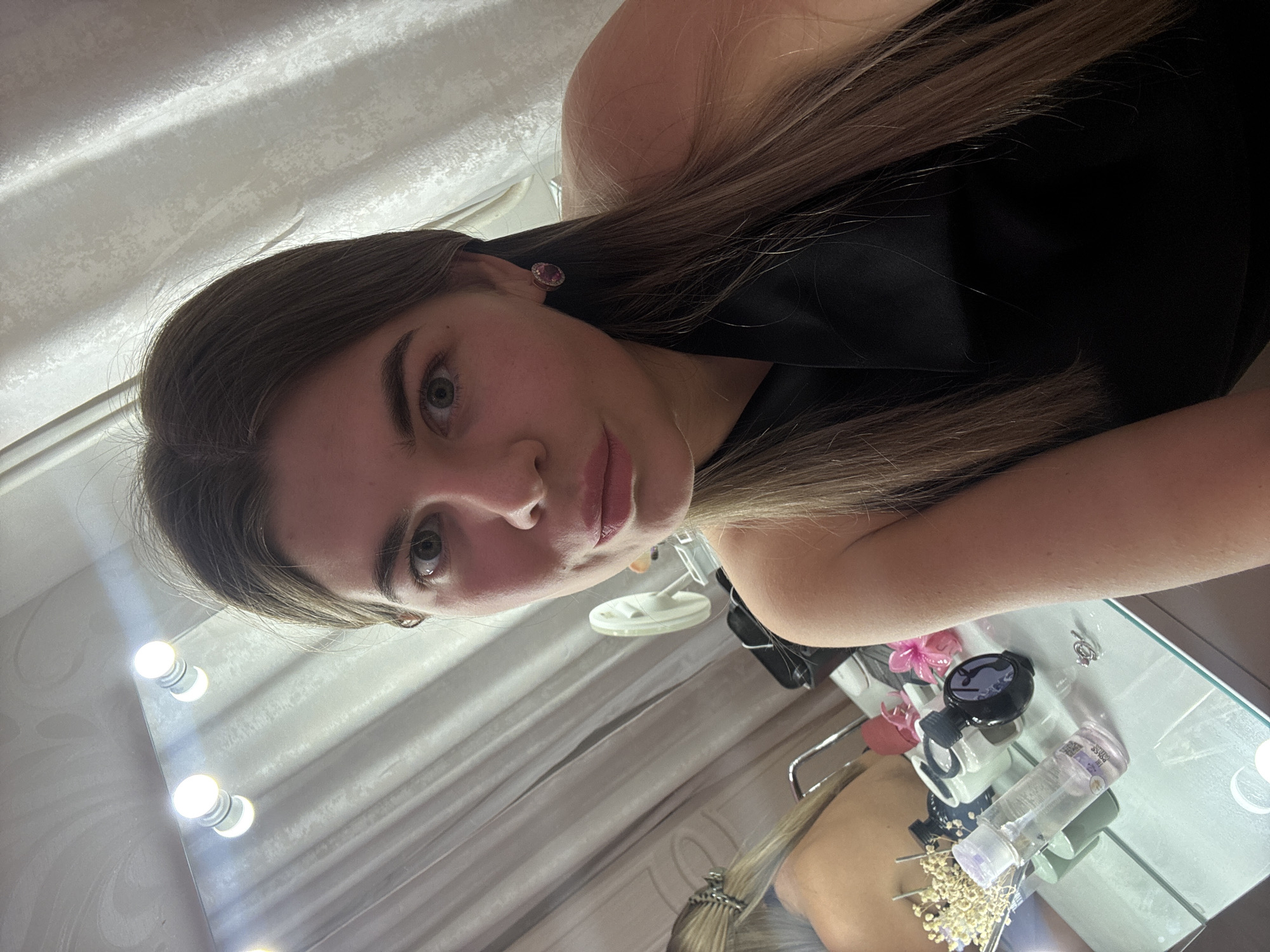 A woman with light brown hair and fair skin is taking a selfie in front of a vanity mirror with illuminated bulbs. She is wearing a black sleeveless top and a pink earring. The vanity table has various beauty products, including bottles and containers. In the reflection, another person is visible. The location is likely a personal residence, potentially a bedroom, as suggested by the vanity and personal items. The exact location within the city of Chaikovsky, Russia, cannot be determined from the image alone.