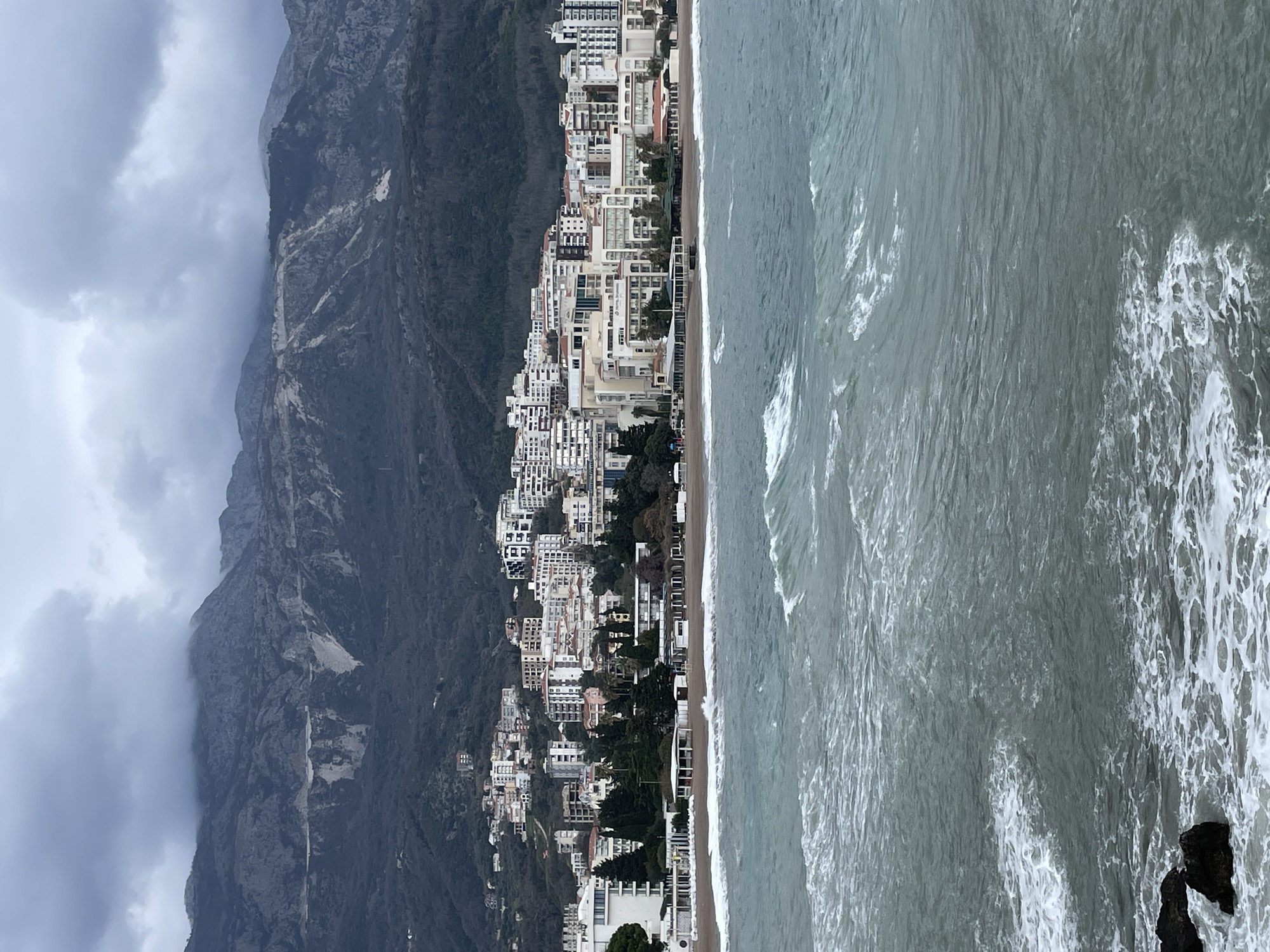 The image captures a wide coastal scene in Budva, Montenegro, specifically depicting the Bečići area under an overcast sky. In the foreground, the grey-blue water of the Adriatic Sea is choppy, with visible white foam from breaking waves. Two dark rock formations are partially submerged in the water at the lower left. A sandy beach, appearing deserted, separates the sea from the urban landscape. Along the coastline and extending up the adjacent hillside, numerous multi-story buildings, identified as hotels and residential structures, are densely constructed. The "Hotel Splendid" sign is clearly visible on one of the prominent beachfront buildings. Various smaller structures, including covered beachfront facilities and white-roofed kiosks, are present on or near the sand. The background features steep, rugged mountains, characterized by dark, sparse vegetation and prominent lighter-colored rock exposures or scars on their slopes. Dense grey clouds obscure the mountain peaks and dominate the sky. No individuals or specific actions are discernible within the frame.