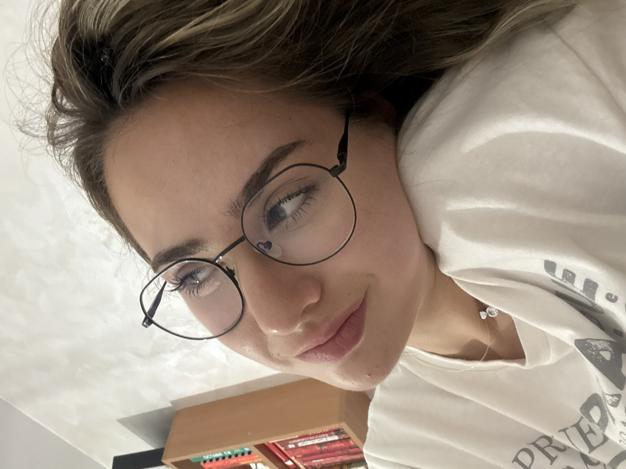 A high-angle selfie captures a young woman, likely taken while she is looking up, with her head resting on a surface. Her face, framed by light brown hair with blonde highlights, dominates the upper part of the image. She wears round, thin-framed dark eyeglasses, through which her light-colored eyes are visible, looking slightly upward and to the side. Her long, dark eyelashes are prominent. She has a subtle, closed-mouth smile or smirk, and her skin appears clear and natural.
She is wearing a white t-shirt, with the partial word "PRIED" visible in dark gray lettering on the fabric near her right shoulder. A delicate silver necklace with a small, clear, possibly floral or butterfly-shaped charm, rests on her collarbone.
The background consists primarily of a plain, light-colored surface, likely a ceiling or wall. In the lower portion of the image, a wooden bookshelf filled with numerous books is visible. Some book spines display text, including Cyrillic characters such as "ВЛАД" (VLAD) and "СТАН" (STAN), while another book appears to have "DISTANT TR..." (possibly "Distant Thunder").
The scene is set indoors, suggesting a home or personal space in Blagoevgrad, Bulgaria, given the location context and visible Cyrillic text. The image conveys a casual and relaxed mood, with the woman appearing contemplative or quietly amused.