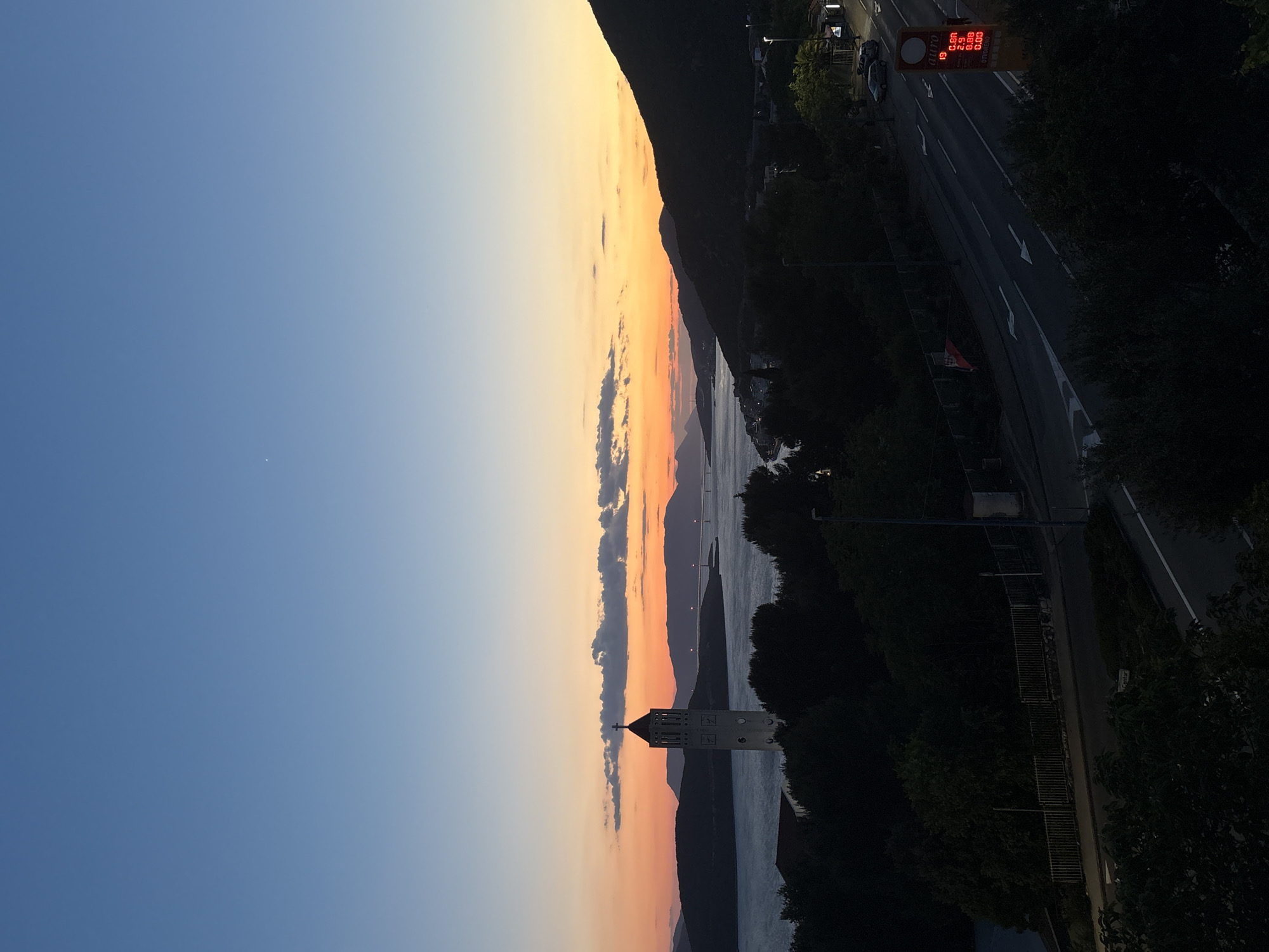 The image depicts a sunset over the Adriatic Sea, likely taken from a higher elevation point in Neum, Bosnia and Herzegovina. To the left, a blue sky fades into orange and yellow hues near the horizon. A line of clouds stretches vertically through the center of the frame, reflecting the sunset colors.
A mountain range runs parallel to the coastline on the right. The sea is visible in the middle, reflecting the sky. A white church tower with a cross at the top stands prominently against the background of the water and mountains.
A road runs along the coastline on the right, with trees lining both sides. A gas price sign is visible on the right side of the road.