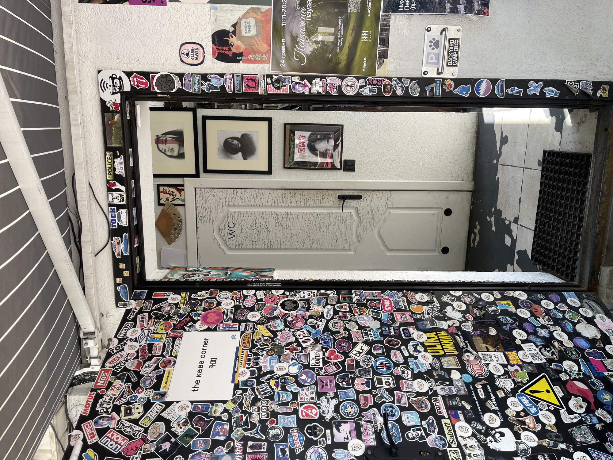 The image captures a vibrant, cluttered entrance or hallway, centered around a large, vertically mounted mirror heavily adorned with an eclectic collection of stickers. The mirror's frame and the wall space around and below it are almost entirely covered in these diverse decals, creating a dense, collage-like surface.
Reflected in the mirror is a white door marked with "WC," indicating a restroom. Beyond this reflected door, an outdoor or semi-outdoor area is visible, featuring a dark floor, possibly concrete, and a black gridded mat near an exit. On the wall above the reflected door, two framed artworks or photographs of faces are hanging.
Above the actual mirror, several posters and flyers are affixed to the white wall. Visible text includes "24 ТРАВНЯ" (May 24), "Пейзажна пауза" (Landscape Break), "TAYONA," "COMING SOON," and "BLACKROLL.UA." A striped, grey and white awning extends downwards on the far left of the frame.
The numerous stickers display a variety of logos, symbols, and brand names, such as "POLICE," "ROCK," a coffee cup with a Wi-Fi symbol, the Rolling Stones logo, the Grateful Dead lightning skull, "Coca-Cola," "Pepsi," "DUNLOP," "Motul," "HRC Honda Racing," "BLUR," and "THE BEATLES." A distinct white sticker below the mirror reads "the kaba corner" and "кафе" (cafe), likely identifying the establishment. A yellow warning triangle sticker with a falling person symbol is also prominent. The scene conveys a quirky, informal atmosphere, characteristic of a cafe, bar, or creative space, under what appears to be daytime lighting conditions.