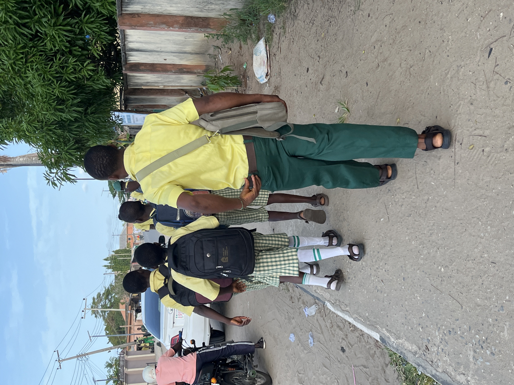 A medium outdoor shot shows a group of people in Badagry, Nigeria. The group consists of four males; one adult, three children. The adult male is wearing a yellow, short-sleeve shirt with a shoulder bag, green pants, and brown sandals. Three children wearing yellow shirts, backpacks, checkered shorts, white socks with green stripes, and dark sandals. They are all walking on a concrete surface, lined with shrubbery and trash. In the background, is a building and light blue sky. Further down the path is a motorcycle, and a white vehicle.