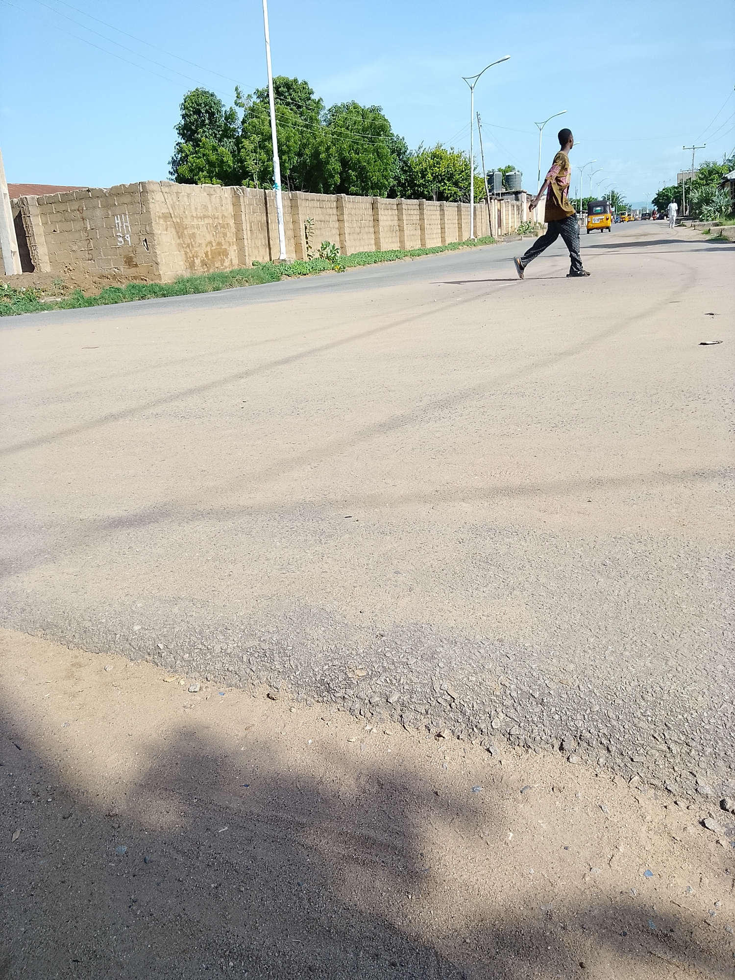 A young man in a patterned shirt and dark pants is walking across a paved road in Wauro Leggal, Nigeria. He is mid-stride, with his right leg forward and his left leg trailing. The road is dusty and appears to be in a semi-arid environment, with a low brick wall and trees lining the left side of the road. In the background, there are several streetlights and some traffic, including a yellow auto-rickshaw and a few other vehicles. The sky is clear and blue, and the sun is casting shadows across the road.