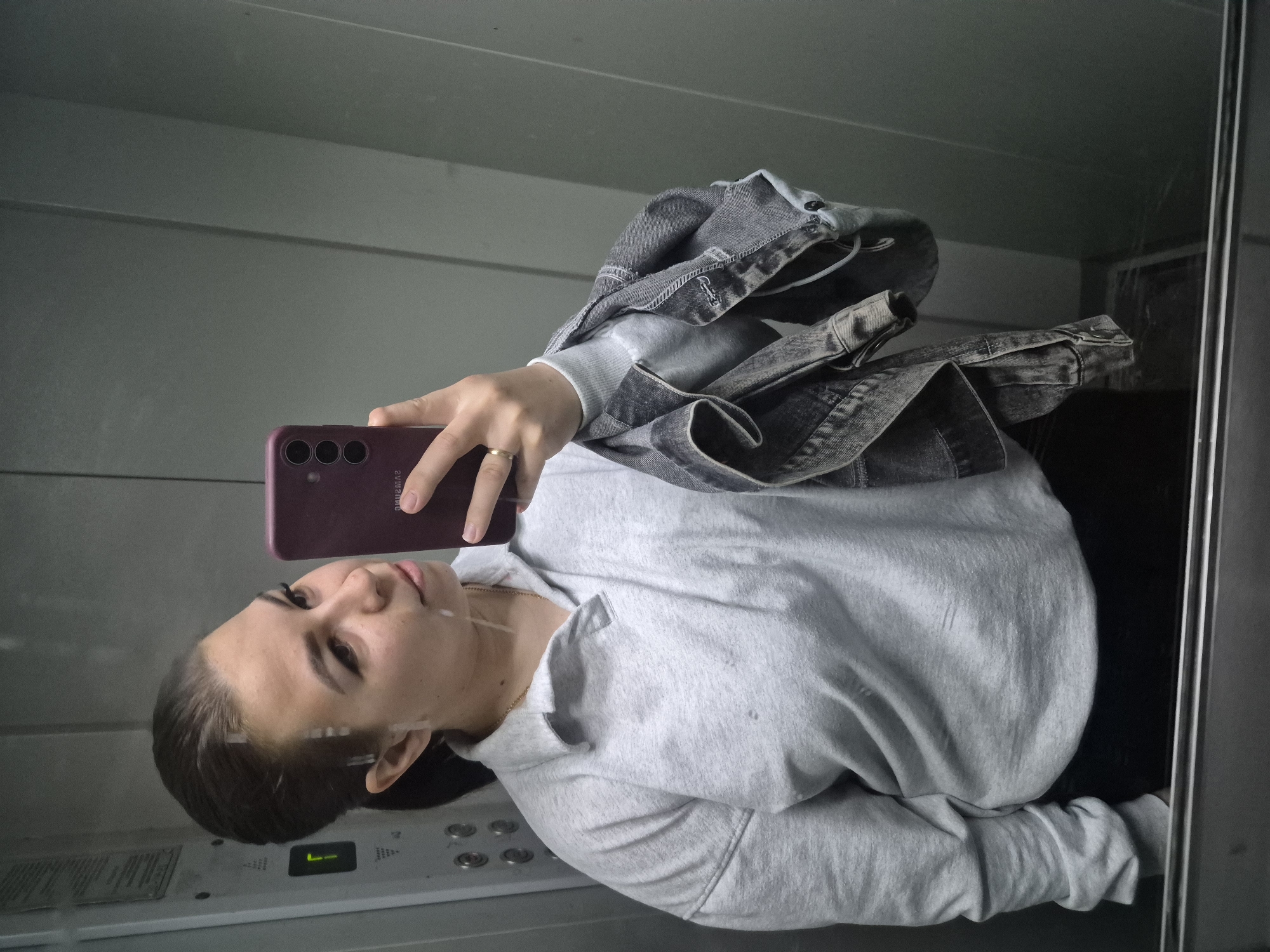 A young woman is taking a selfie in an elevator. She is holding a maroon-colored smartphone with three rear cameras. She is wearing a light grey hooded sweatshirt and a denim jacket is draped over her shoulder. Her hair is pulled back, and she is wearing a ring on her finger. The elevator has a panel with buttons and an indicator light. The elevator appears to be in Tobolsk, Russia.