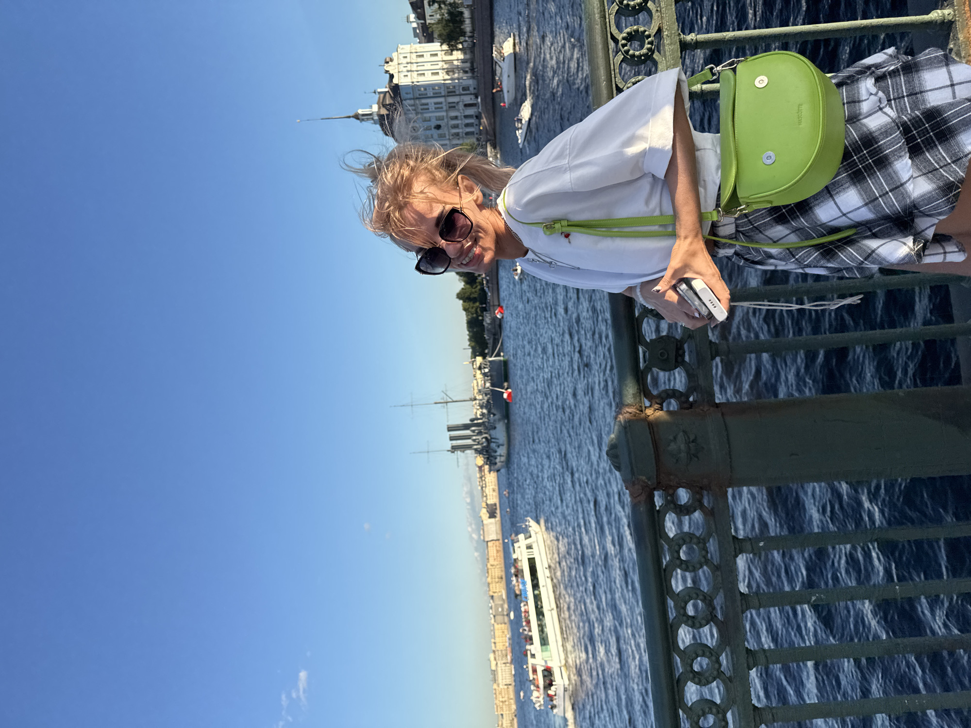 A young woman with blonde hair pulled back in a ponytail, wearing large sunglasses, a white t-shirt, and a black and white plaid skirt, stands on a bridge railing. She holds a white smartphone in her right hand and a bright green cross-body purse with a flap closure hangs from her left shoulder. In the background, a wide blue river flows, with several boats visible on the water. On the far bank of the river, a row of European-style buildings with varied architecture line the waterfront, including a large white building with a spire. The sky is clear and blue, indicating a sunny day. The railing of the bridge is dark green metal with an ornate, circular design.