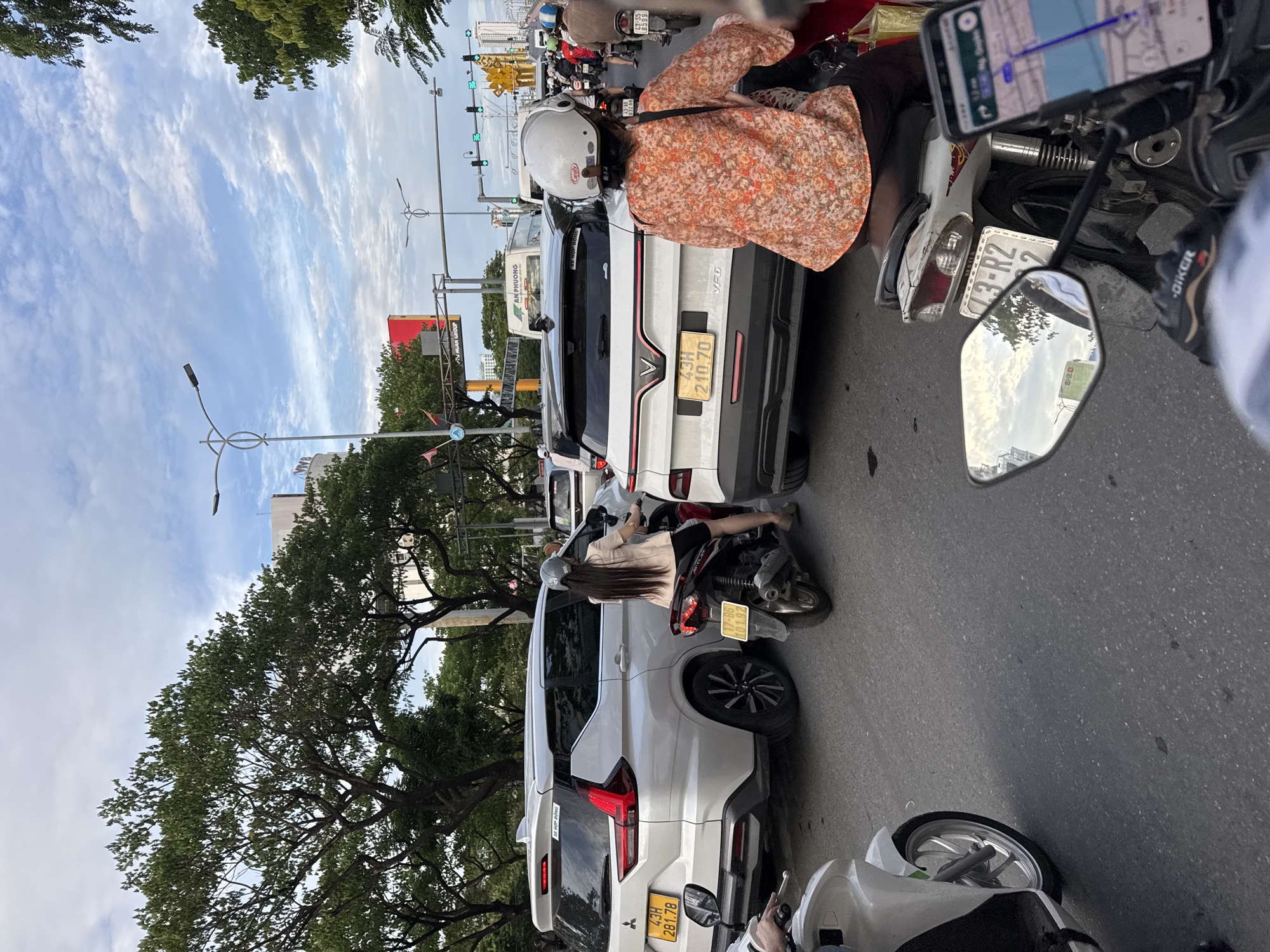The image depicts a traffic jam in Da Nang, Vietnam. Multiple vehicles, including cars and motorcycles, are stopped on a paved road under a cloudy sky. Trees line the street, and traffic lights are visible in the distance. A white SUV is prominent in the foreground, with a person on a motorcycle to its right. Another motorcycle is partially visible on the left. The scene appears to be captured from the perspective of someone on a motorcycle.