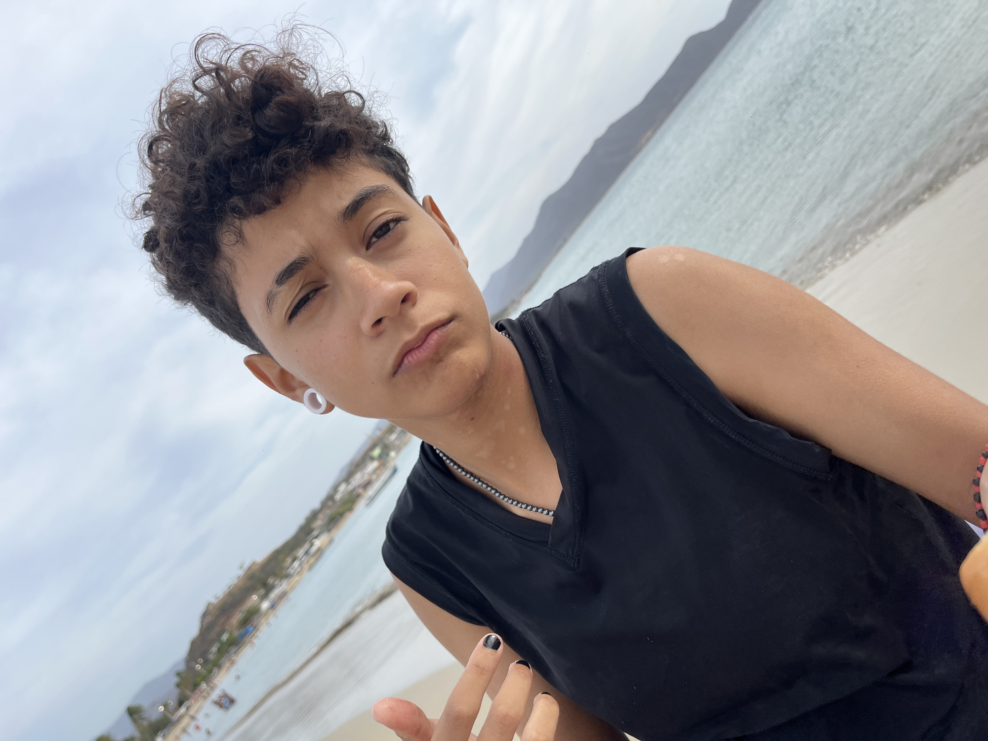 A young person with curly, dark hair is posing for a selfie. They are winking, wearing a black sleeveless top and a beaded necklace. Their fingernails are painted black. The background shows a beach with the ocean and a city skyline, including a prominent hill. Based on the location clues given, this image appears to have been taken in Juan Griego, Venezuela, along the shoreline. The sky is overcast.