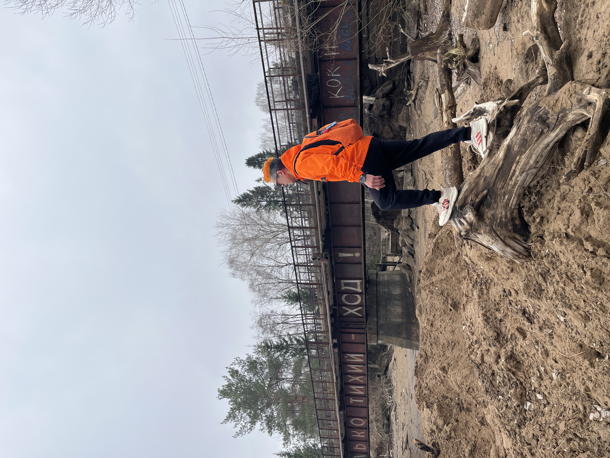 The image shows a person standing on a tree root on the bank of a body of water in Chaikovsky, Russia. The person is wearing an orange jacket, black pants, white sneakers with red stripes, and a cap that matches the jacket. They have orange hair. A backpack is visible on their back.
Behind the person is a metal bridge with Russian text painted on it. The text is "ЛЬКО ТИХИЙ - ХОД!", and "КОК" is also visible. Trees and a gray sky are in the background. The riverbank appears to be made of sandy soil.