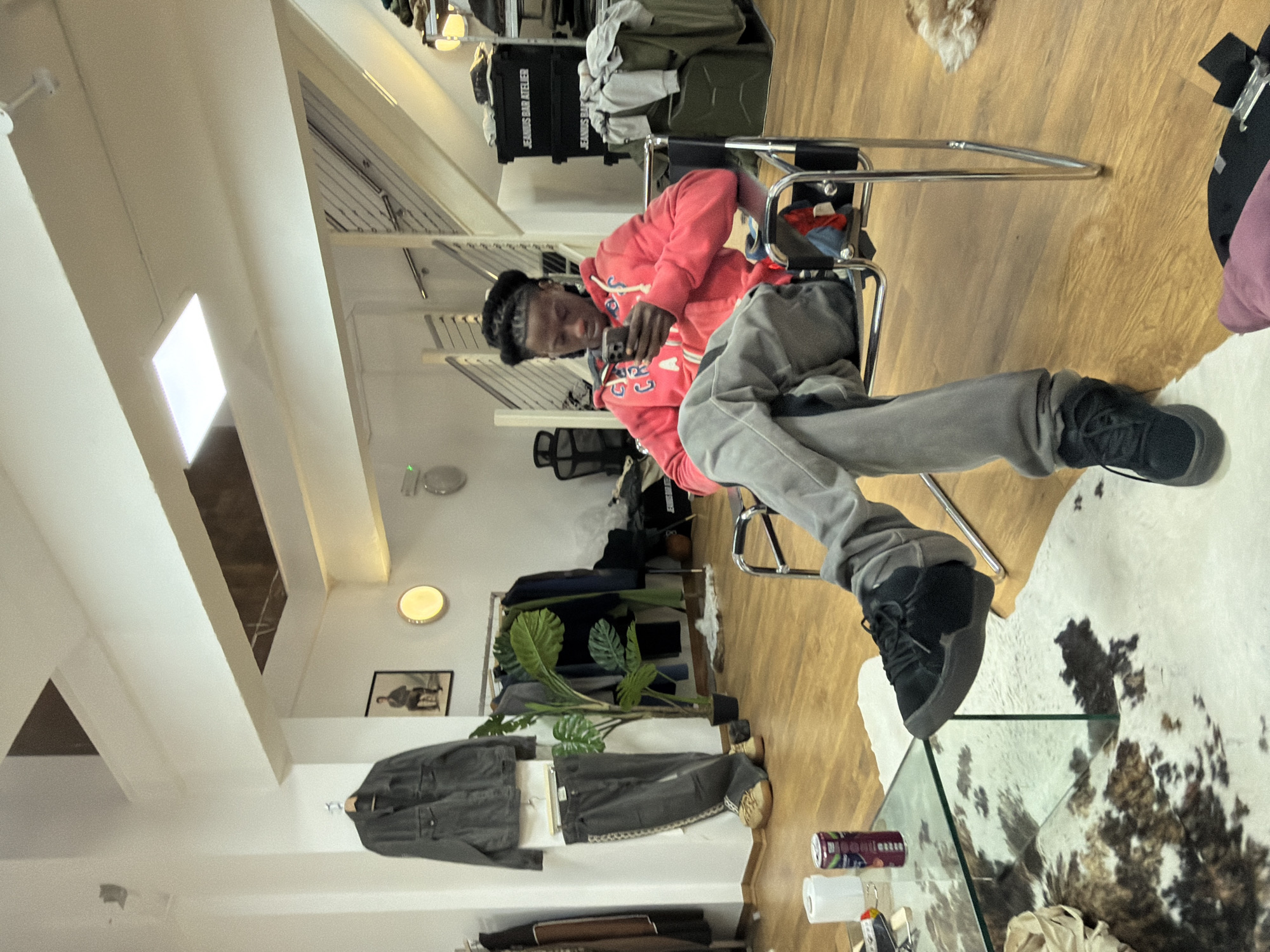 The scene is set inside the Jeansuit Bar Atelier, a retail establishment located in London, United Kingdom. A young adult Black male is seated centrally in a modern chair with a chrome frame and black fabric. He wears a red hooded sweatshirt with white lettering, light grey trousers, and black lace-up sneakers, with his right foot resting over his left knee. He is focused on a dark-colored smartphone held in both hands. In the immediate foreground, a glass-topped coffee table displays a white paper roll and a purple canned beverage. A white and brown cowhide-patterned rug partially covers the light-colored wooden floor beneath the table.
To the left, a dark grey denim jacket and matching trousers hang on a wall display above two pairs of tan shoes. An artificial green plant in a black pot stands nearby, adjacent to a clothing rack laden with various garments. The background features a modern staircase with metal railings ascending to an upper level. Beneath the staircase, several dark green or black industrial-style containers are stacked, with "JEANSUIT BAR ATELIER" printed in white text on their sides, and textile items piled on top. The walls are white, and the ceiling is equipped with recessed square lights and a circular wall-mounted light.