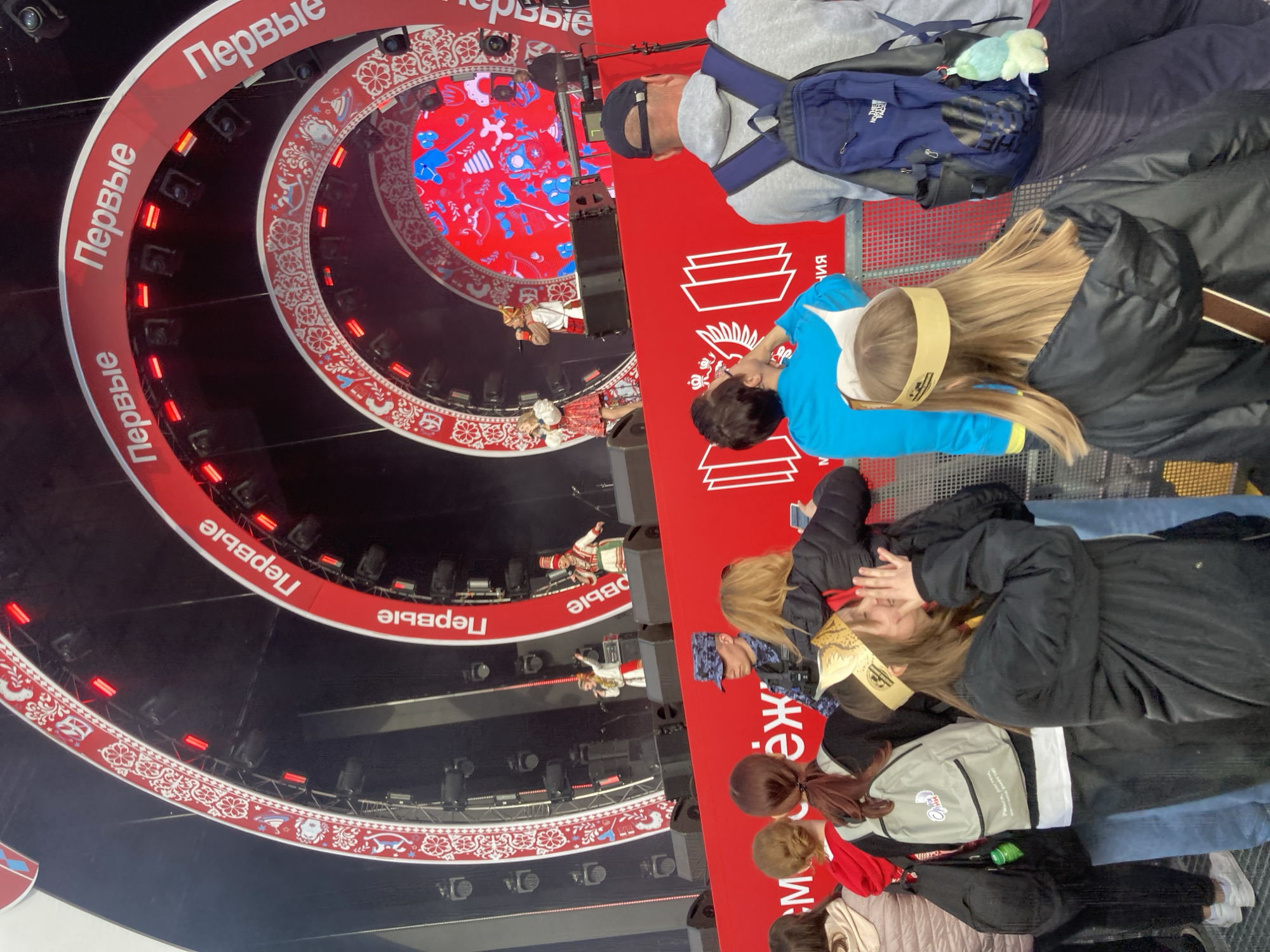 The image shows an event taking place in Moscow, Russia, as evidenced by the Cyrillic script "Первые" (Pervyye) which translates to "The First" and the presence of traditional Russian patterns and costumes on stage. The setting appears to be a large arena or indoor venue designed with concentric circular platforms. A performance is underway, with performers in traditional Russian attire. A small audience is visible in the foreground, consisting of adults and children who are observing the performance, some holding phones. The image shows the children wearing headbands with the event name on it. A man with a blue backpack, and a black cap is also visible in the audience. The backdrop of the stage features a screen displaying red, white and blue graphics.
