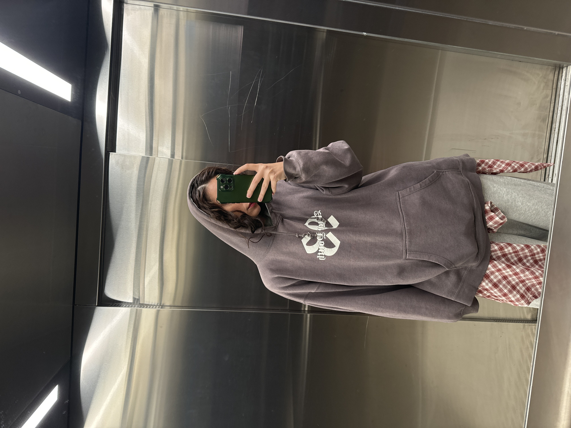 A young woman is taking a selfie in what appears to be an elevator with stainless steel walls. She is wearing a purple hoodie with a white design on the front, grey sweatpants, and a red and white checkered shirt underneath the hoodie. She is holding a green iPhone. The lighting in the elevator is bright. The specific location in Krasnoyarsk, Russia cannot be determined from the image alone.