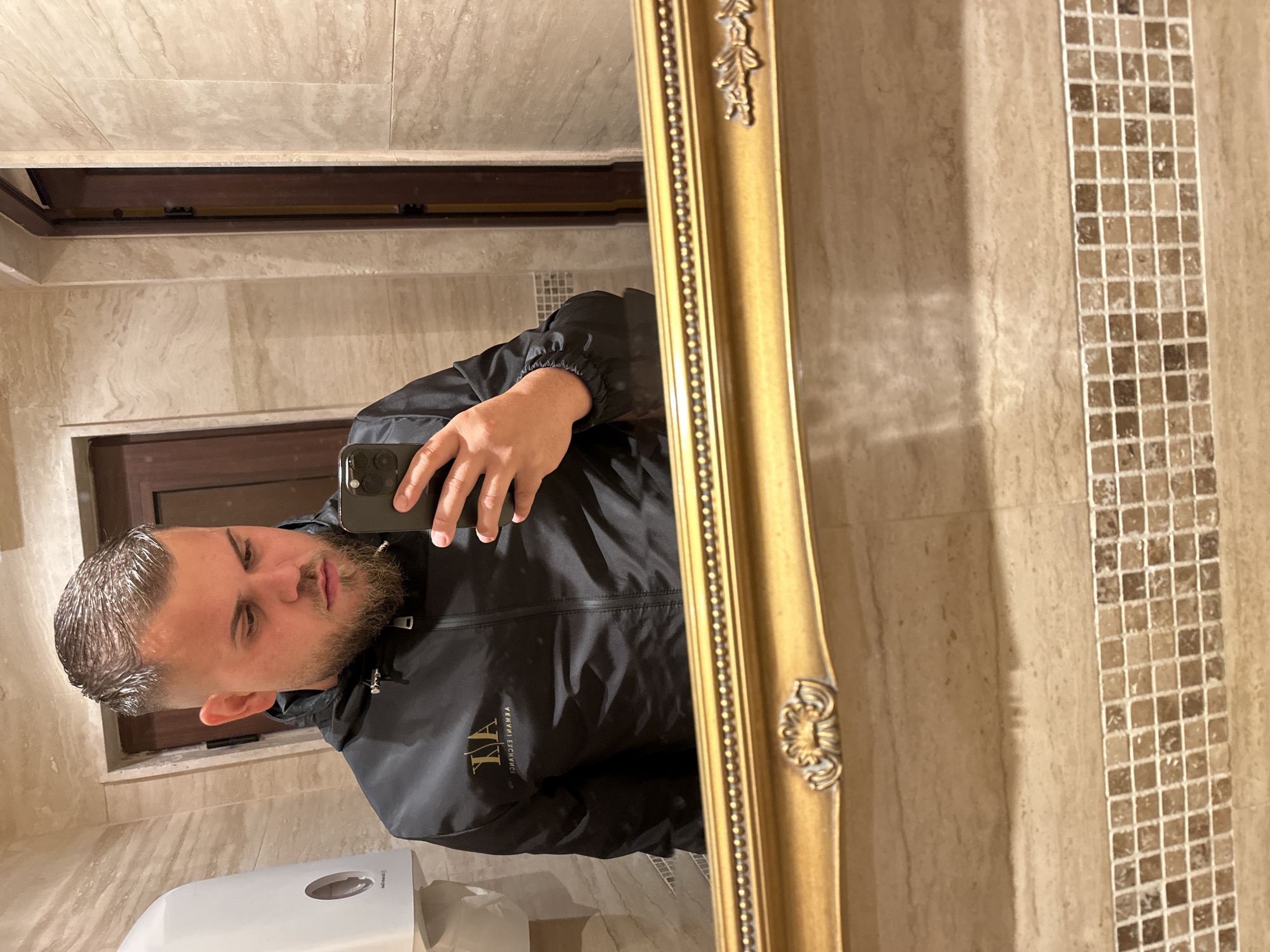 A young man with short, styled blonde hair and a beard takes a selfie in a bathroom. He is wearing a black zippered jacket with a gold logo on the chest. The bathroom features beige tiled walls and a gold-framed mirror. A white soap dispenser is visible on the left. The scene appears to be in a public restroom within the city of Sozopol, Bulgaria, possibly in a restaurant or hotel.