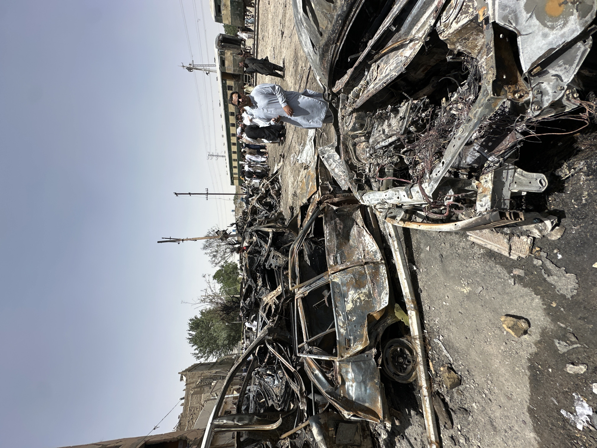 The scene depicts extensive damage to vehicles, reduced to charred and twisted metal, indicative of a potential explosion or severe fire. A group of men, several wearing traditional attire, stand observing the scene. One man in a light-colored shalwar kameez appears to be inspecting the wreckage closely. The scene appears to be located in the city of Quetta City Tehsil, Pakistan, close to railcars. Power lines and utility poles are visible in the background. The overall environment suggests an aftermath of violence or accidental disaster.