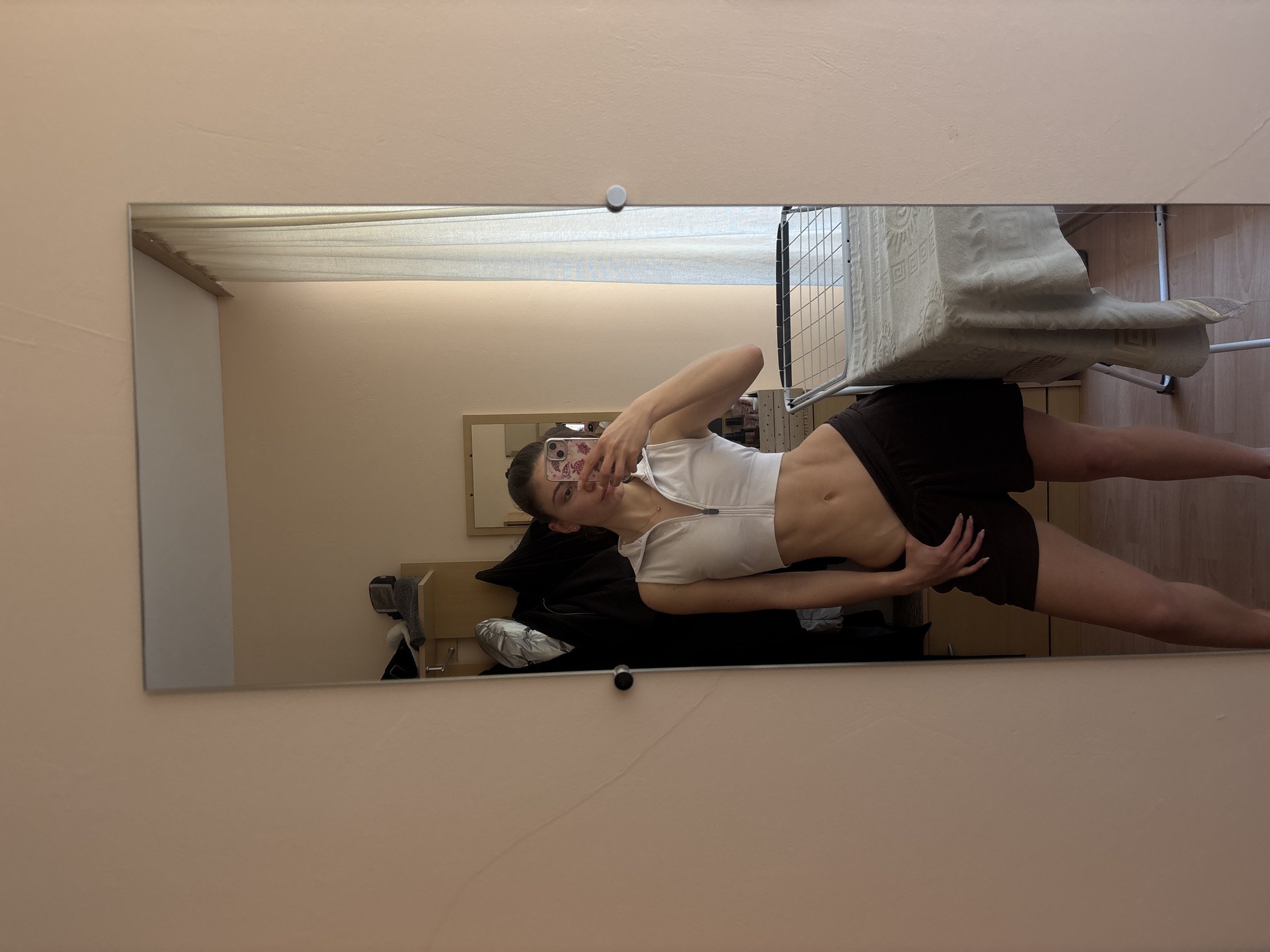 The image, rotated 90 degrees counter-clockwise, captures a young woman taking a mirror selfie in an indoor setting, likely in Varna, Bulgaria. The subject, appearing to be in her late teens or early twenties with dark hair styled back, is wearing a white, short-sleeved, zipped crop top and dark shorts, showcasing her midriff. She holds a smartphone with a patterned case up to her face to take the photo, while posing with her left hand placed on her hip or lower back.
The large rectangular mirror reflects a room with light, warm neutral-colored walls. Visible behind the woman's reflection is a small, light-colored framed mirror or picture on the wall. To the right of her reflection (viewer's left), a wooden piece of furniture, possibly a headboard, is seen, along with what appears to be a towel or fabric and a coat rack with hanging items. To her left (viewer's right), a metal drying rack holds a folded, patterned blanket or rug. The floor in the reflection is light-colored wood or laminate.
Two white circular clips, one at the top and one near the bottom, secure the mirror to the wall. The room appears well-lit. No text is visible in the image. The woman has a slender physique and light-colored, manicured fingernails.