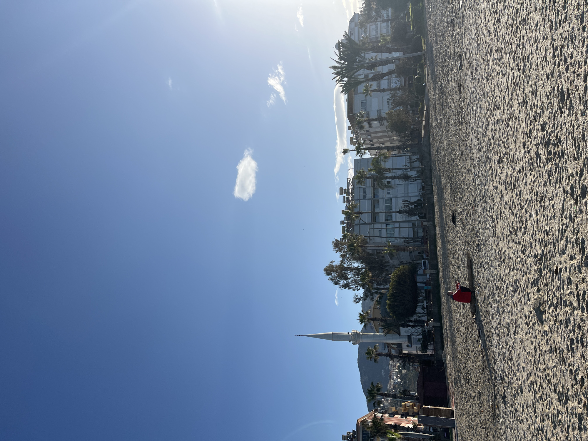 The image depicts a beach scene in Alanya, Turkey, under a clear, blue sky with a few scattered clouds. The right side of the frame is dominated by a sandy beach, and a single person is lying on the sand, wearing a red jacket. The person appears to be sunbathing or resting.
On the left side of the frame, there's a view of the city. Buildings, including what appears to be a mosque with a tall minaret, are visible, suggesting a populated area. Palm trees line the streets, typical of the Mediterranean environment. A mountain is visible in the background. The city seems to be positioned between the beach and the mountainous terrain.