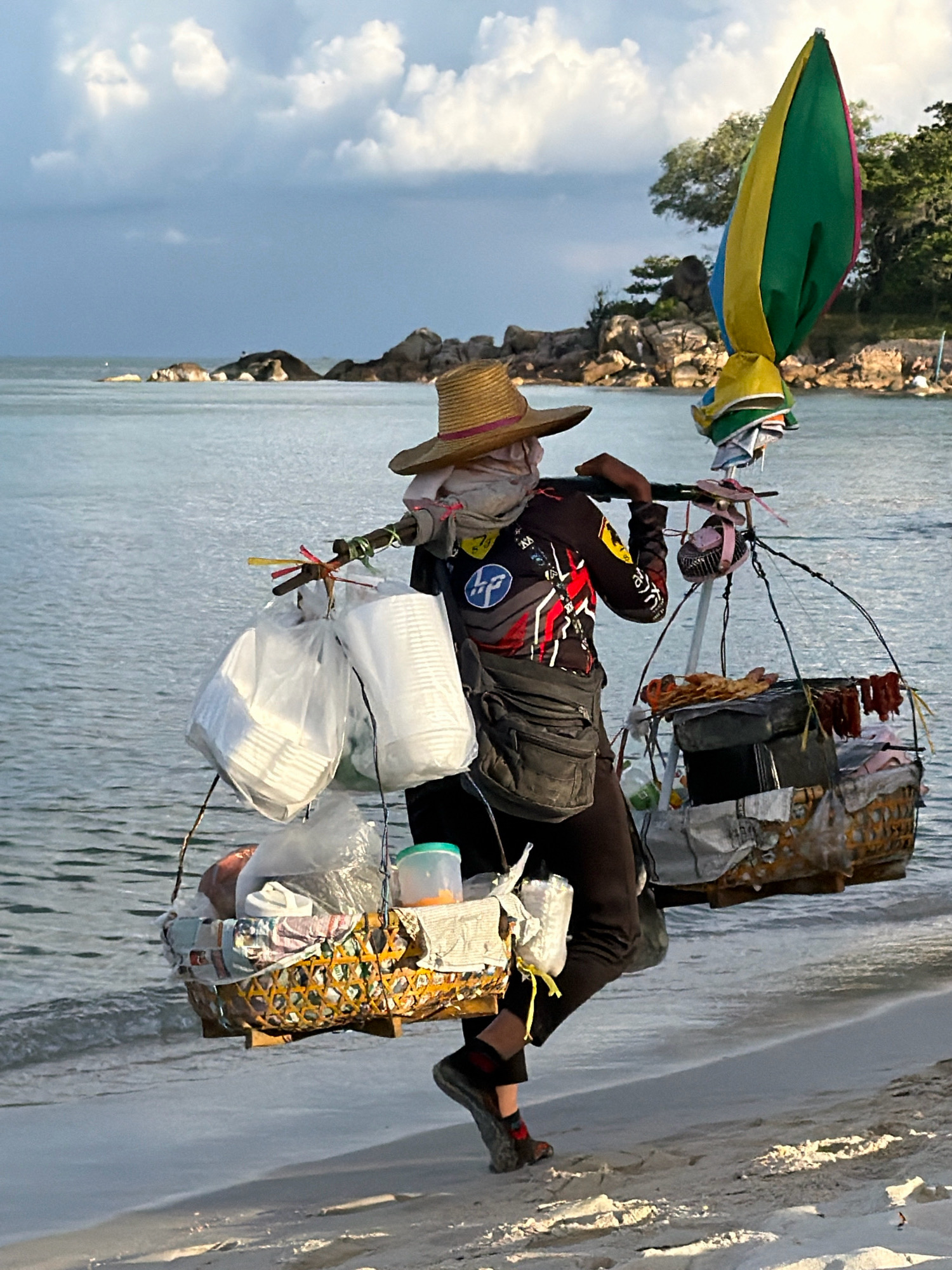 A person wearing a straw hat and a long-sleeved shirt with a design is walking on a sandy beach. They are carrying a large load on their shoulders using a pole. The load consists of several baskets and bags, containing what appears to be food items, plastic containers, and other goods. A colorful umbrella is attached to the top of the load. The background shows the sea with calm water and some rocky outcrops, as well as trees on the distant shore, indicative of a tropical coastal environment. The lighting suggests it is either early morning or late afternoon, with soft light illuminating the scene. The person's posture and the arrangement of the load suggest they are a vendor. The presence of the sea, sand, and tropical vegetation points to a location like Ko Samui District, Thailand.