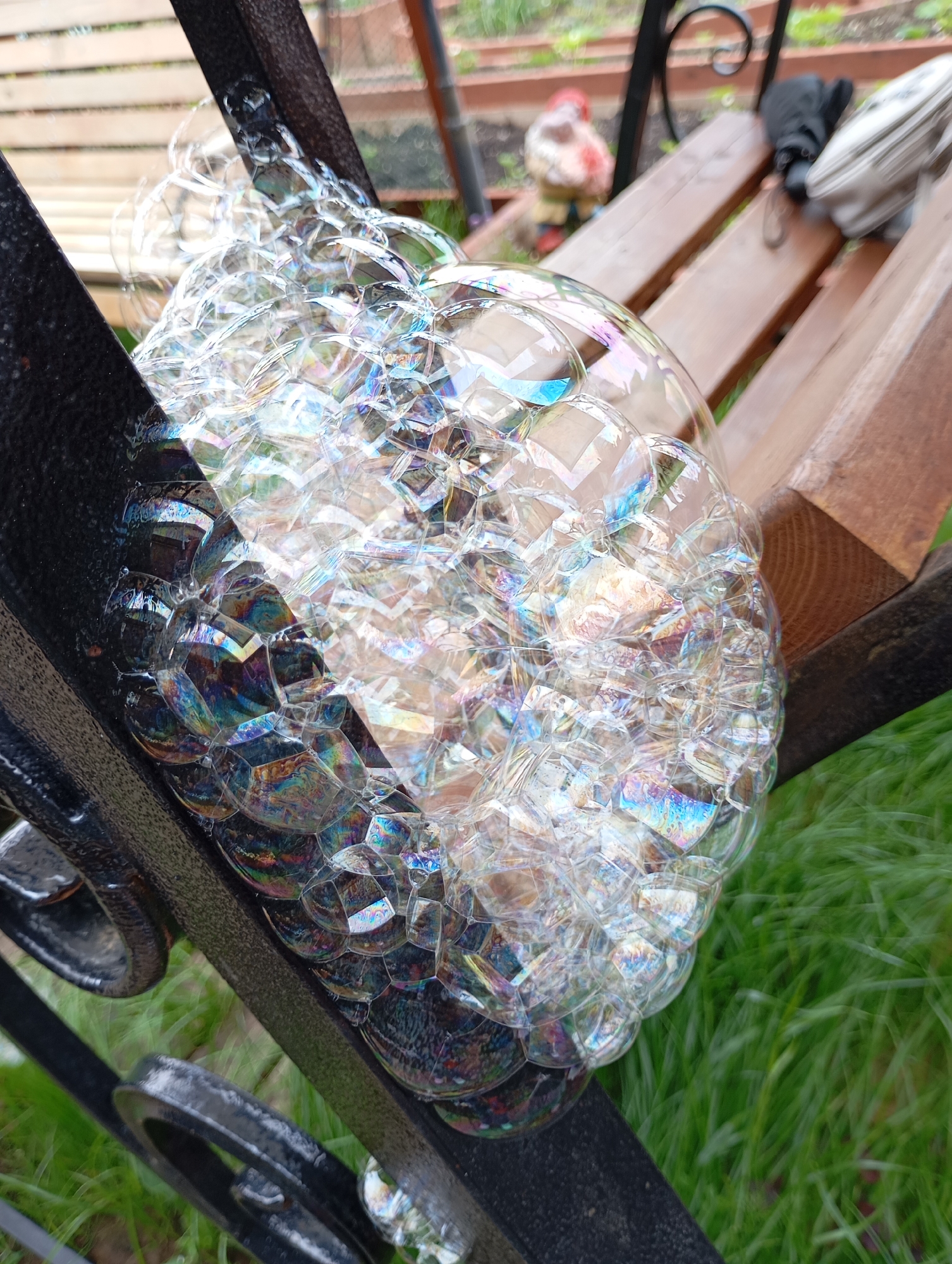 A cluster of iridescent soap bubbles rests upon a dark-colored metal structure, possibly a fence or railing, positioned diagonally across the lower and central portions of the frame. The bubbles exhibit a spectrum of colors including blues, purples, greens, and yellows due to light interference. The metal structure features a textured, matte finish and includes visible curved and straight elements. In the blurred background, several horizontal light-brown wooden slats, consistent with a bench or decking, are visible. To the left of the wooden slats, an out-of-focus garden figurine, possibly a gnome with red and white features, is discernible. On the rightmost wooden slat, a light-colored bag or object with a dark strap is partially visible. Below the metal structure and wooden elements, vibrant green grass is present, mostly out of focus. The scene is located in Фоки, Russia.