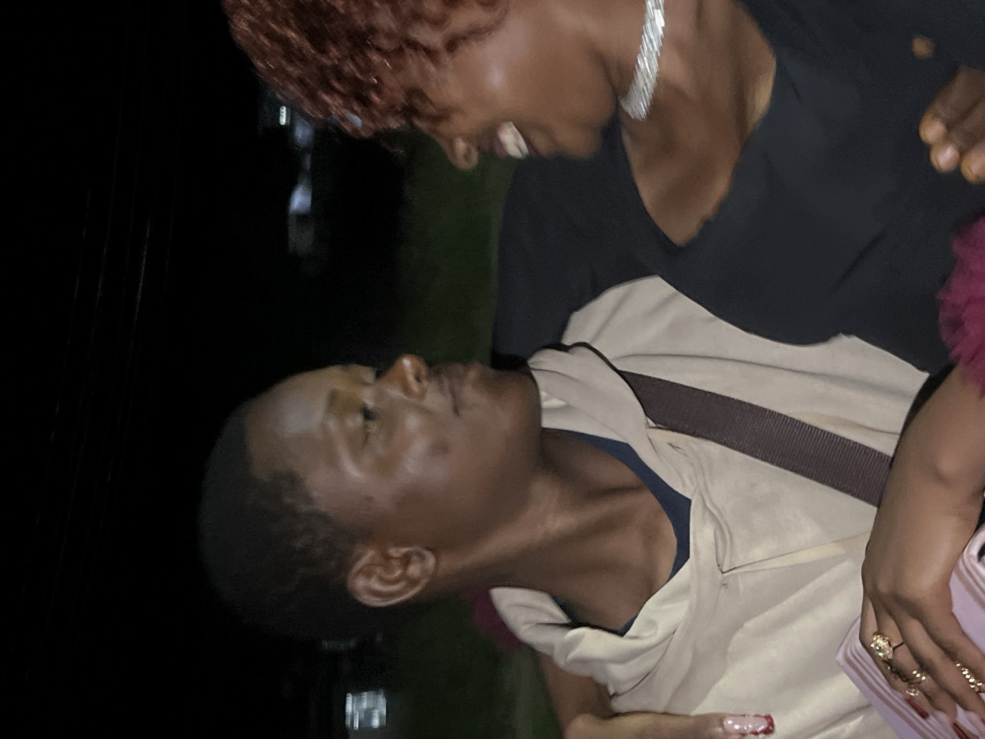 Two individuals are pictured at night, with the woman on the right laughing and looking at the man on the left. The man is wearing a light-colored hooded sweatshirt with a blue undershirt and a brown strap across his chest. The woman is wearing a black top and a sparkling necklace. The background appears to be an outdoor urban environment with some indistinct lights and foliage visible. The city of Durumi, Nigeria is mentioned, suggesting this scene could be taking place there.