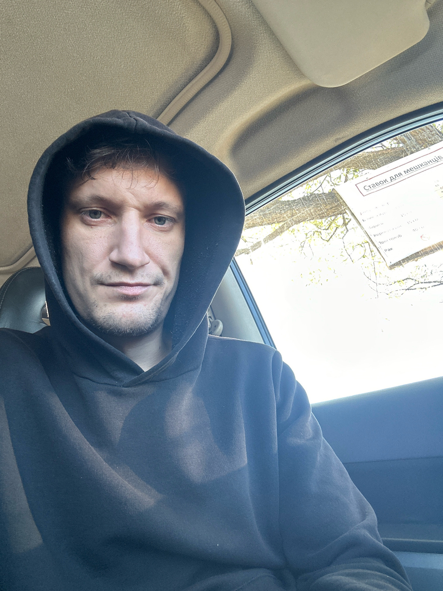 A person, likely male, is seated inside a vehicle, facing the camera and wearing a dark hooded sweatshirt. The individual has light skin, short brown hair, and stubble. The interior of the vehicle appears to be a car, with a beige headliner and a sun visor visible. Through the car's window, which has some condensation on it, a bright sky and the branches of trees are visible, suggesting it is daytime. A white sign with text in Cyrillic is affixed to the window. The lighting suggests natural daylight is coming from the right side of the frame.