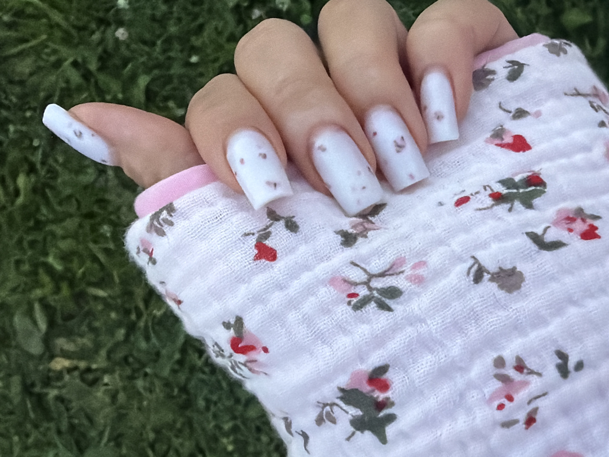 The image shows a close-up of a person's hand and fingernails, against a blurred green background that appears to be grass. The hand is partially covered by a white cloth with a floral pattern of pink and red flowers and green leaves. The fingernails are long, square-shaped, and painted white with small brown speckles. The thumb, index finger, middle finger, and ring finger are visible. The image does not contain any recognizable landmarks or features that would allow for identification of a specific location within the city of Красный Маяк, Russia.