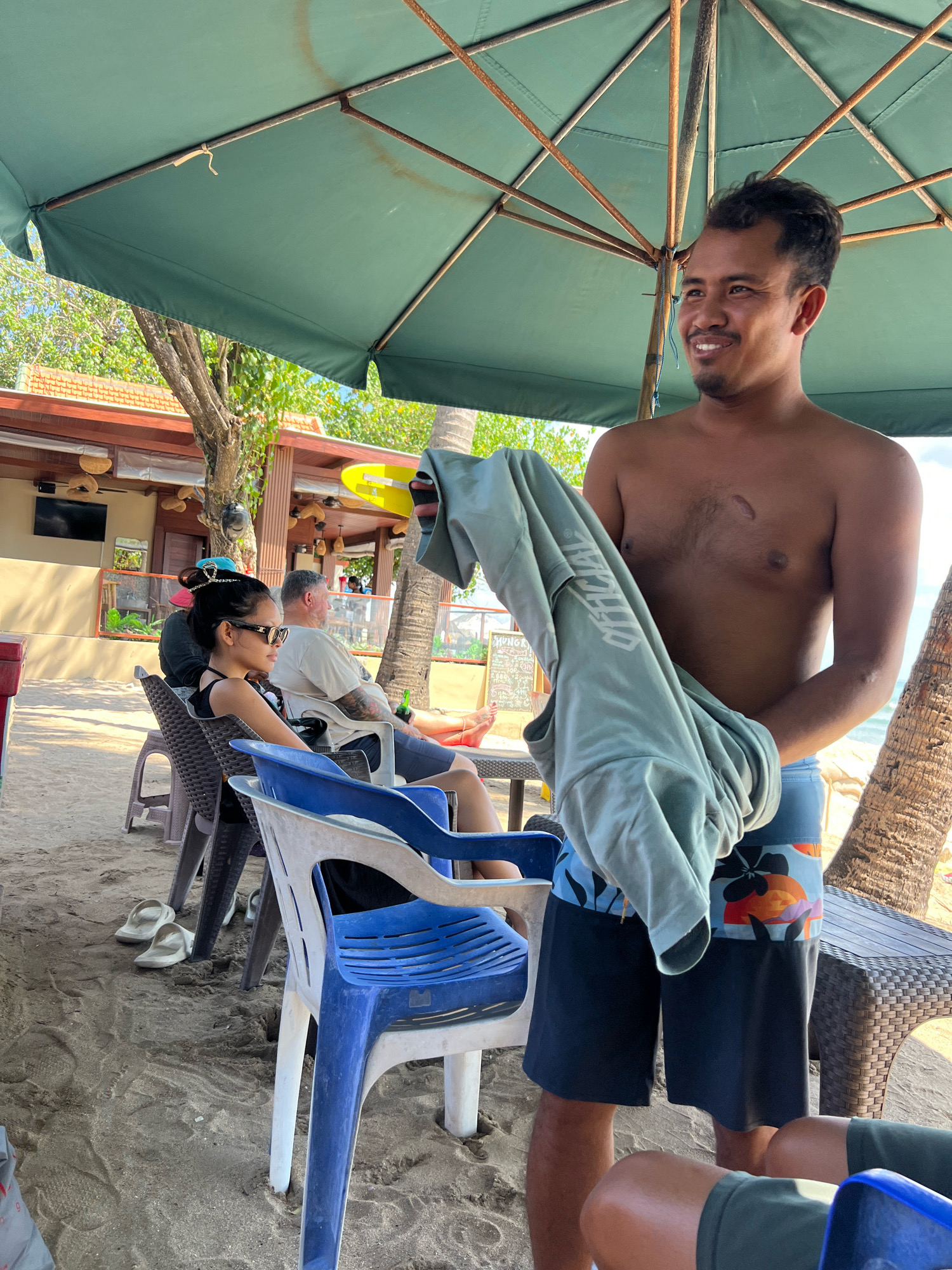 A shirtless man with dark hair and a smile stands in the foreground, holding a light grey t-shirt. He is wearing patterned swim shorts. Behind him, people are seated at tables under a large green umbrella on a sandy beach. In the background, a building with a red roof and outdoor seating is visible, with palm trees and the ocean beyond. The scene appears to be a beachside establishment in Kuta, Indonesia.