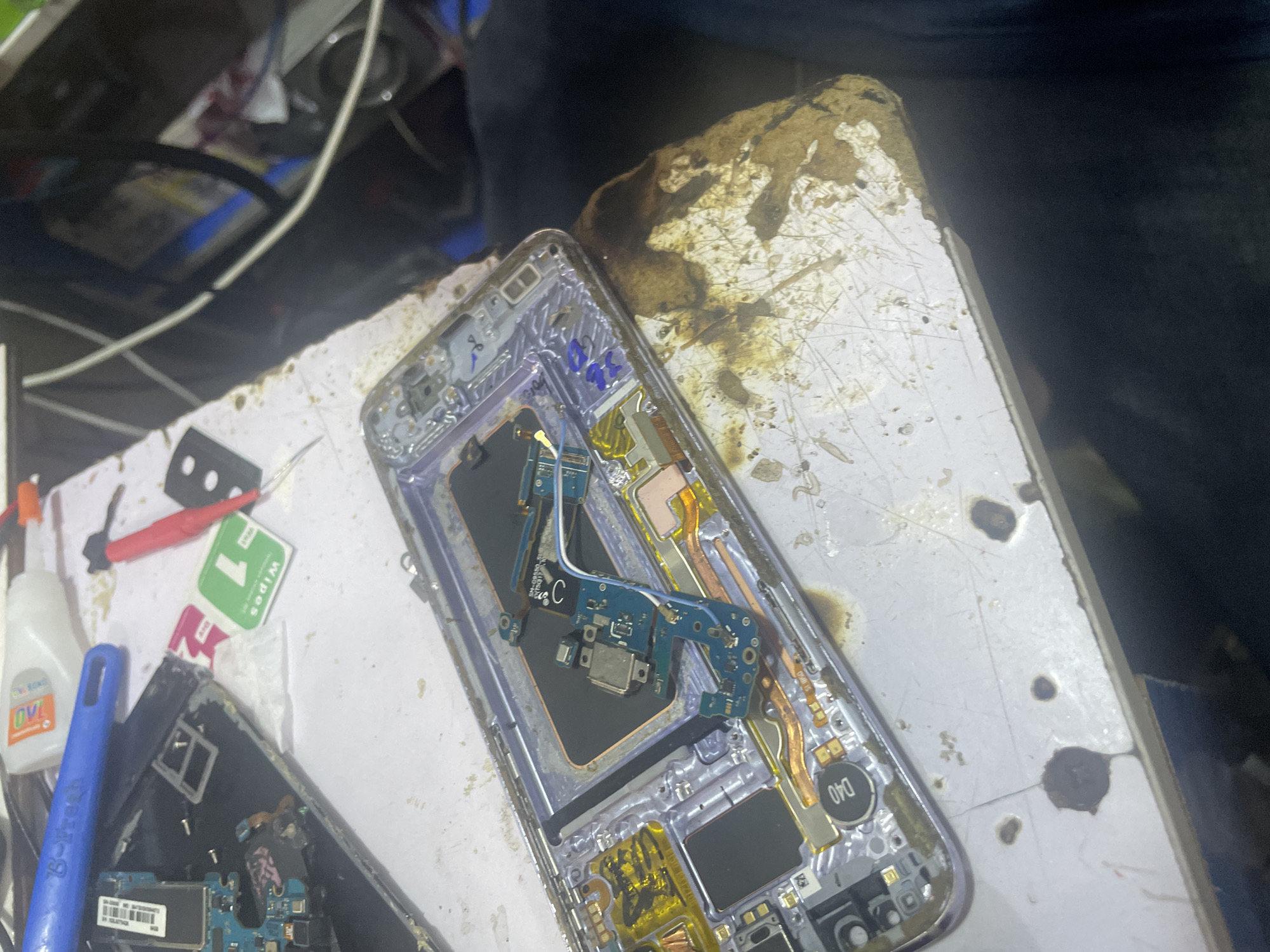 The image depicts a close-up, overhead view of a disassembled smartphone laid out on a cluttered workbench. The phone's internal components, including the circuit board, battery, and charging port, are visible. The workbench surface is stained and appears to be a workspace for electronics repair, with tools such as a blue pry tool and a small red screwdriver lying nearby. A white bottle with orange lettering, likely containing adhesive or cleaning fluid, is also present. A green packet labeled "WIPES 1" is visible, suggesting a cleaning process is underway or has been completed. The overall scene is one of technical work, likely phone repair or maintenance. The background is dark and out of focus, making it difficult to discern further details of the environment. There are some dark, burnt-looking stains on the workbench, hinting at past soldering or heat-related work. The lighting suggests an indoor setting, possibly a workshop or repair shop, with no clear indicators of time of day or weather. The focus is entirely on the electronic components and the immediate workspace.