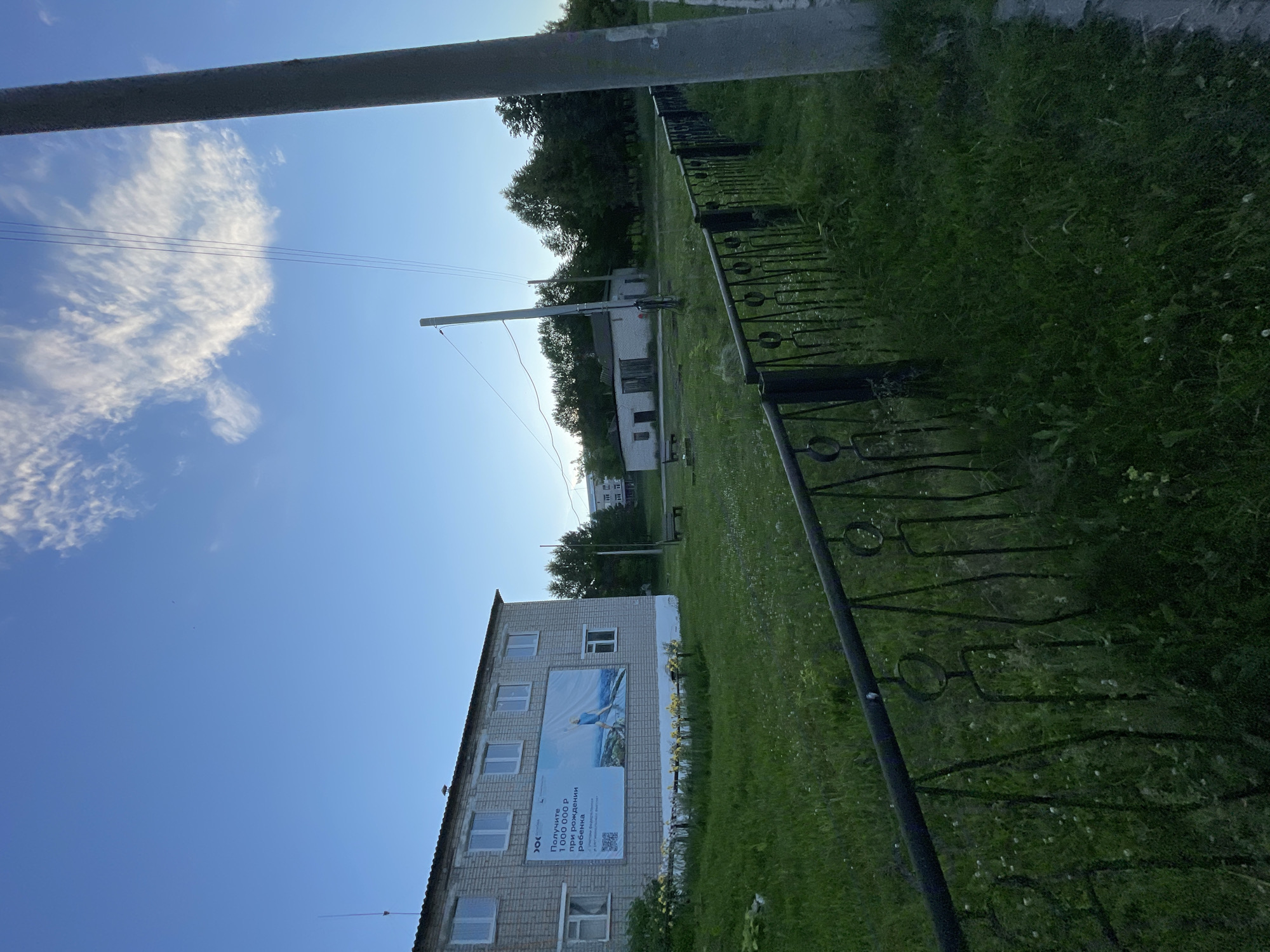 The image depicts a residential building in Krasnye Baki, Russia, under a clear sky with scattered clouds. The building, constructed of light-colored brick, features multiple windows and a large billboard on its facade. The billboard shows an image of a person standing on a mountaintop and Russian text. In front of the building, a patch of grass is dotted with small white flowers. A black metal fence with decorative circular elements runs along the foreground. In the background, there are trees, another building, and utility poles with wires. The overall scene suggests a rural or suburban setting during daylight hours.