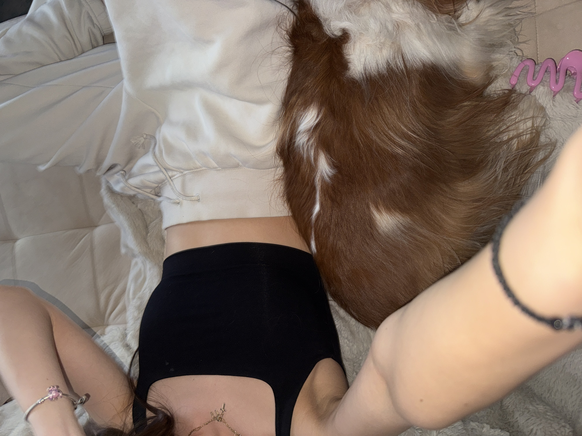 An individual, appearing to be female, is lying partially supine on a light-colored, plush, and tufted surface, likely a bed or sofa, in an indoor setting. They are wearing a black sleeveless crop top and light beige or off-white sweatpants. A silver-toned bracelet with a prominent pink floral charm and a smaller heart-shaped charm is visible on their left wrist. A delicate gold-toned necklace adorned with multiple small charms, including a horseshoe and a cross, rests on their chest. Dark hair is visible near the left shoulder.
To the right of the individual, a small to medium-sized long-haired dog with distinctive reddish-brown and white fur patches is resting on the same surface, positioned alongside the individual's torso. The dog's fur is predominantly reddish-brown with significant white markings, particularly on its back and flanks. A pink, wavy, plastic object, possibly a hair accessory or toy, is partially visible on the surface near the dog's rear. The lighting is soft and ambient. The image provides no visual indicators to confirm a specific location within Sofia, Bulgaria.