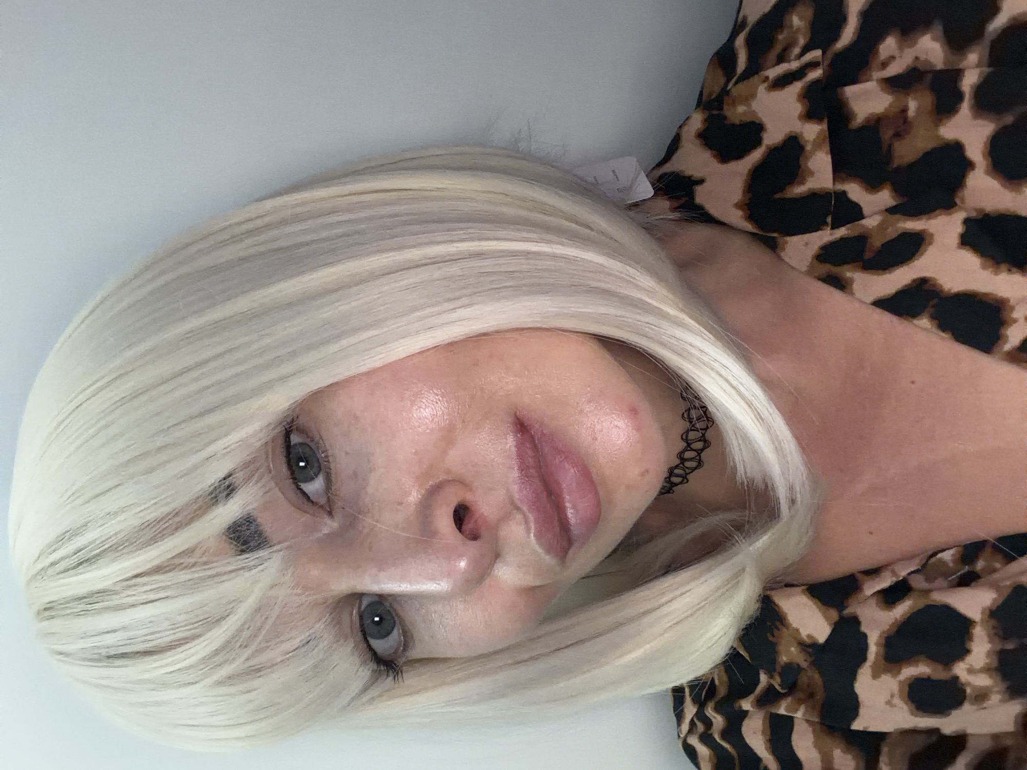 A close-up portrait features an individual with light blonde, bob-length hair and straight bangs covering the forehead. The individual has light blue-grey eyes, which gaze directly forward, and dark, defined eyebrows visible through the fringe. The skin exhibits a dewy texture with visible pores, minor freckling on the cheeks, and a few small blemishes, notably on the chin and near the nasal area. The nose is prominent, and the lips are full, appearing light pink with a subtle sheen.
The individual wears a collared garment with a brown and black leopard print pattern. A black, intricate, patterned choker necklace is worn around the neck. The background consists of a plain, light-colored wall, indicating an indoor setting. A small white tag or piece of paper is partially visible behind the hair on the right side of the individual's neck. The subject's head is slightly tilted, presenting a direct, neutral expression.
The image provides no discernible visual information to determine a specific geographical location, such as the city of Saint Petersburg, Russia, or any other city.