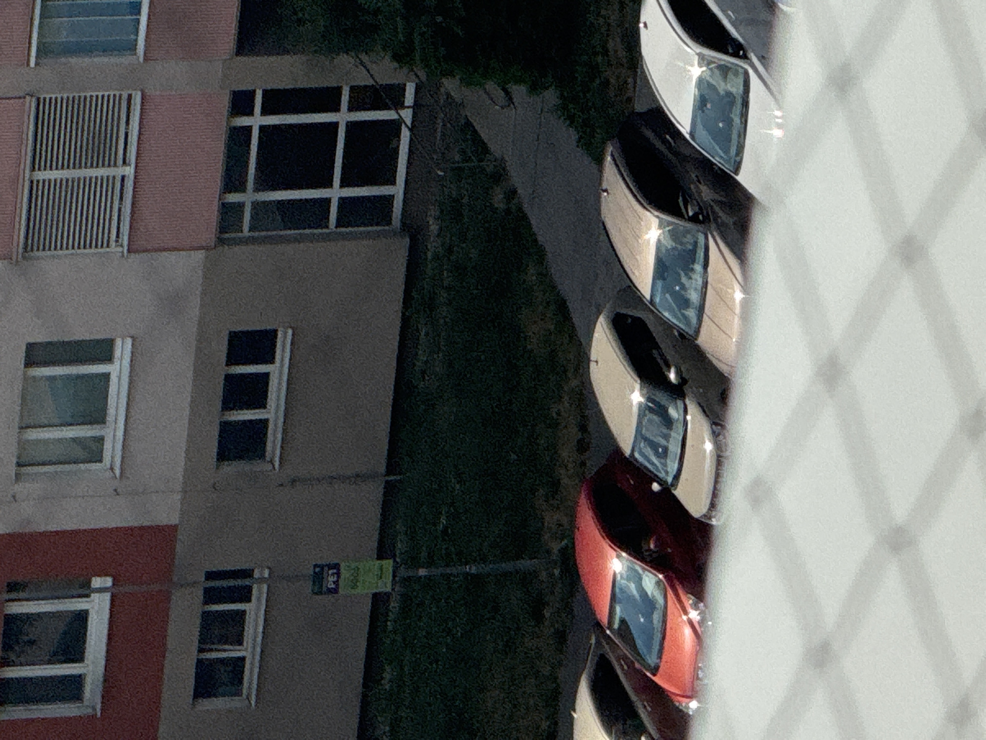 The image captures a scene in Bratislava, Slovakia, focusing on a residential building with parked cars. The building features a reddish-brown facade on the upper left, transitioning to a grey lower section. Several windows are visible, some with horizontal blinds. In front of the building, a row of cars is parked on a paved area adjacent to a grassy slope. The cars, from left to right, include a dark vehicle, a bright red hatchback, a beige/tan SUV, and a white SUV. A pole with two signs is also visible, one blue with white text that appears to say "PET" and a green sign below it with white text and numbers. The foreground is dominated by a blurred, white, grid-like structure, suggesting the image was taken through a fence or similar barrier. The overall impression is that of an urban residential environment on a sunny day.