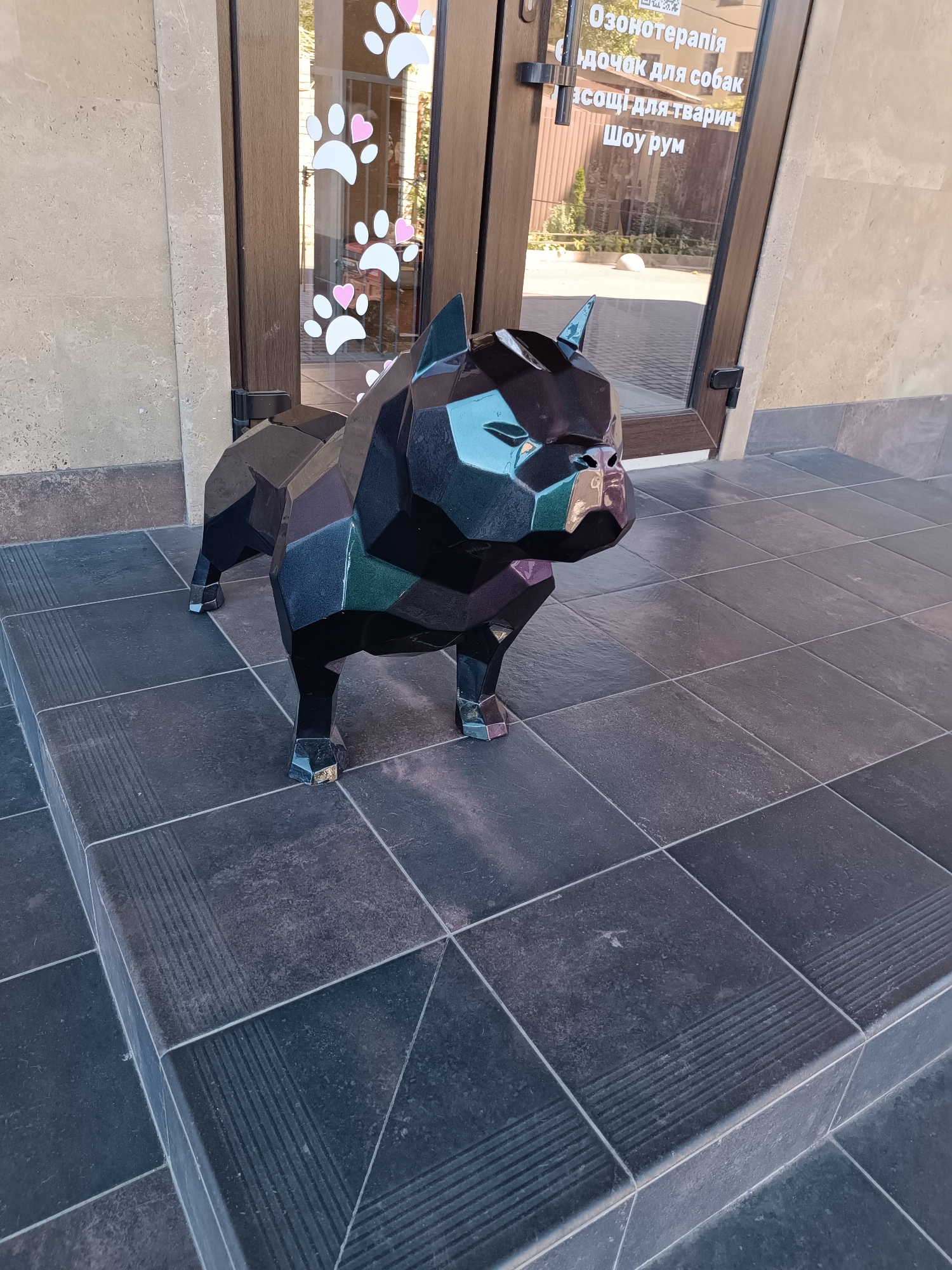 The image shows a faceted black sculpture of a dog standing on a tiled outdoor staircase. The sculpture has a stylized, geometric design with sharp angles and varied reflective surfaces that catch the light, showing hints of purple and green. The dog sculpture is positioned in front of a glass door, which is decorated with white paw prints and pink hearts. Text is visible on the glass door, appearing to be in Ukrainian. To the left of the door, there is a stone wall. The ground and stairs are tiled with dark gray, rectangular tiles. The overall scene suggests the entrance to a commercial establishment, possibly a pet-related business.
