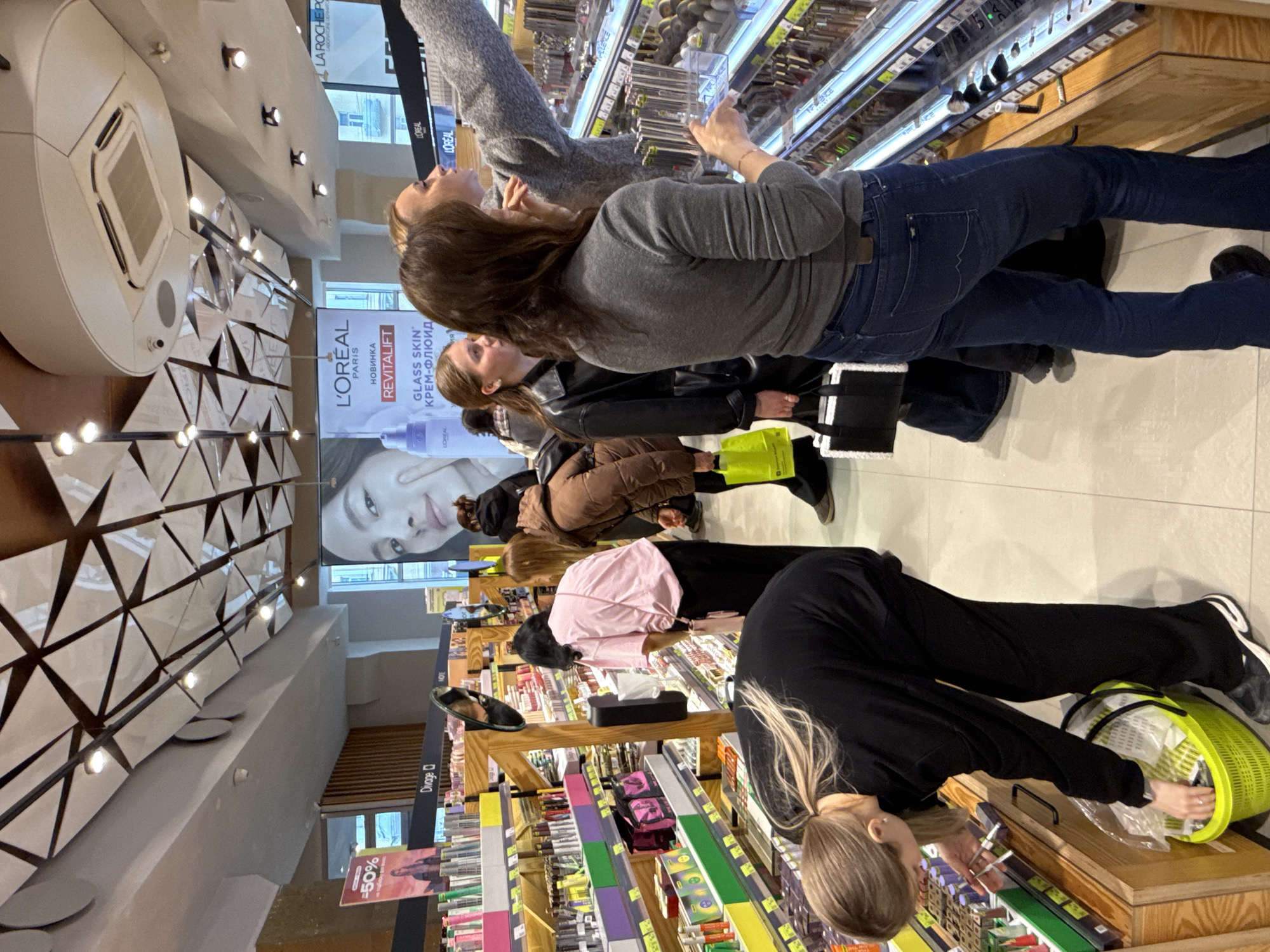 Here is a description of the image:
The image shows the interior of a store, likely a cosmetics or beauty supply store, in Kazan, Russia. Several women are browsing the shelves of makeup and skincare products. They are reaching out and examining the items. There is a sign displaying "L'ORÉAL PARIS" along with Russian text "НОВИНКА REVITALIFT GLASS SKIN' КРЕМ-ФЛЮИД", indicating it is an advertisement for a L'Oréal product. Other visible brands include "Divage" and "LA ROCHE-POSAY". One of the women is using a lime green basket.