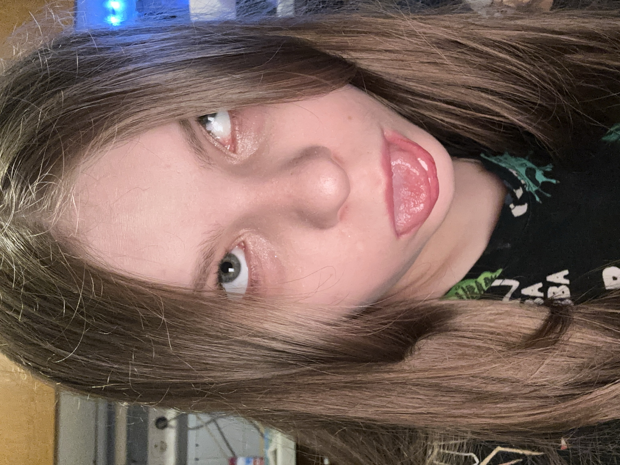 A close-up, slightly angled, indoor portrait shows a person with light skin and long, light brown hair. The person has grayish-green eyes and is sticking out their tongue. They are wearing a dark-colored shirt with a green and white pattern. The background is blurry, showing a mix of light-colored walls and indistinct objects. The location cannot be determined from this image alone. There is no discernible evidence to suggest it is in Moscow, Russia.