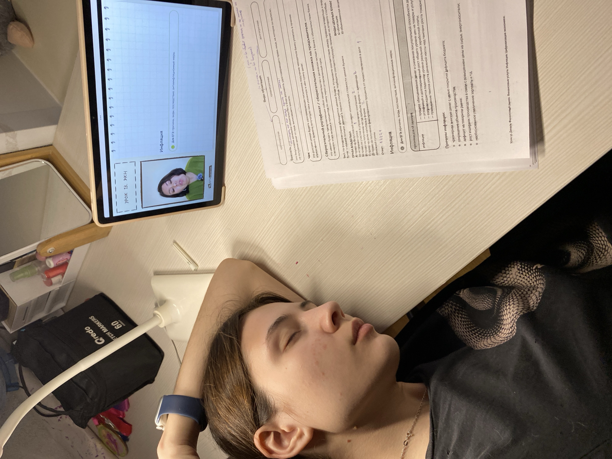 A young woman is asleep at a desk, her head resting on her left hand. An open book, titled "Инфляция" (Inflation), lies next to her. A tablet showing a lesson titled "УРОК 13. ЭКН" (Lesson 13. EKN) is propped up nearby. There is also a desk lamp. There are also markers and pens. The scene is indoors. The location is not discernible from the image.