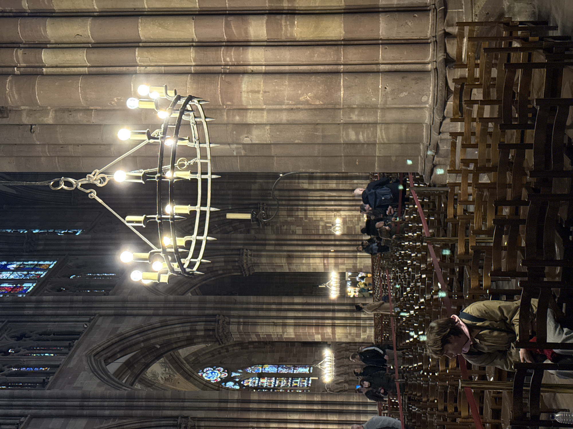 The image displays the interior of Cathédrale Notre-Dame de Strasbourg, France, focusing on a section of the nave or a main aisle. Prominent features include towering sandstone columns with a reddish-brown hue, pointed arches, and vibrant stained glass windows on the upper left, primarily depicting blue and red patterns. A large, ornate circular metal chandelier, equipped with multiple electric light bulbs designed to resemble candles, hangs in the upper right quadrant, illuminating the immediate area. Several smaller, similar chandeliers are visible further into the cathedral.
The floor area is densely populated with rows of dark wooden chairs, arranged for seating. Red ropes are strung between posts, delineating pathways and seating sections.
In the immediate foreground, a person with brown hair, wearing a tan jacket over a pink hoodie and light-colored pants, is seated on one of the wooden chairs, facing left and slightly upwards. A dark backpack is situated behind them, and a clear plastic water bottle is visible on the floor to their left. In the midground and background, approximately fifteen to twenty other individuals are present. They are primarily standing or slowly moving, observing the cathedral's interior. Notable individuals include a person in a dark jacket with a blue backpack near a column in the midground and another individual in a long tan coat standing further back, facing away from the camera. The overall lighting is subdued, with primary illumination originating from the suspended chandeliers and filtered light from the stained glass.