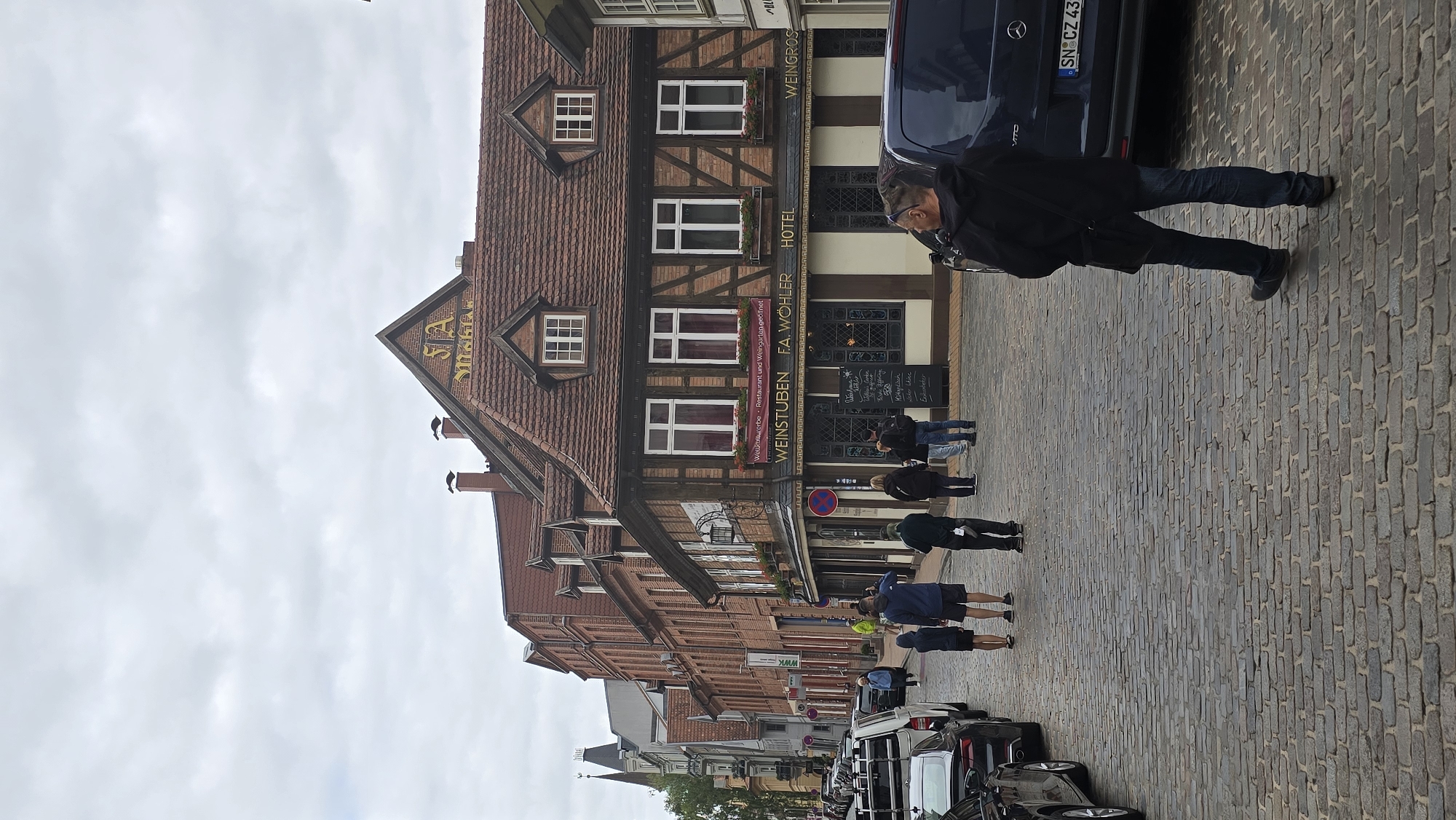 This image shows a street scene in Schwerin, Germany. The central focus is a historic building with a red tiled roof, timber framing, and multiple windows. The building displays signage indicating "WEINSTUBEN F.A. WÖHLER HOTEL" and "WEINGROSS", suggesting it is a wine tavern, hotel, and wine shop. The street is paved with cobblestones. Several people are present in the scene: a man in a black jacket and jeans is walking away from the camera, a group of people are standing on the sidewalk in front of the building, and a few individuals are walking down the street. Cars are parked on the left side of the street and a dark blue van is parked on the right. The sky is overcast.