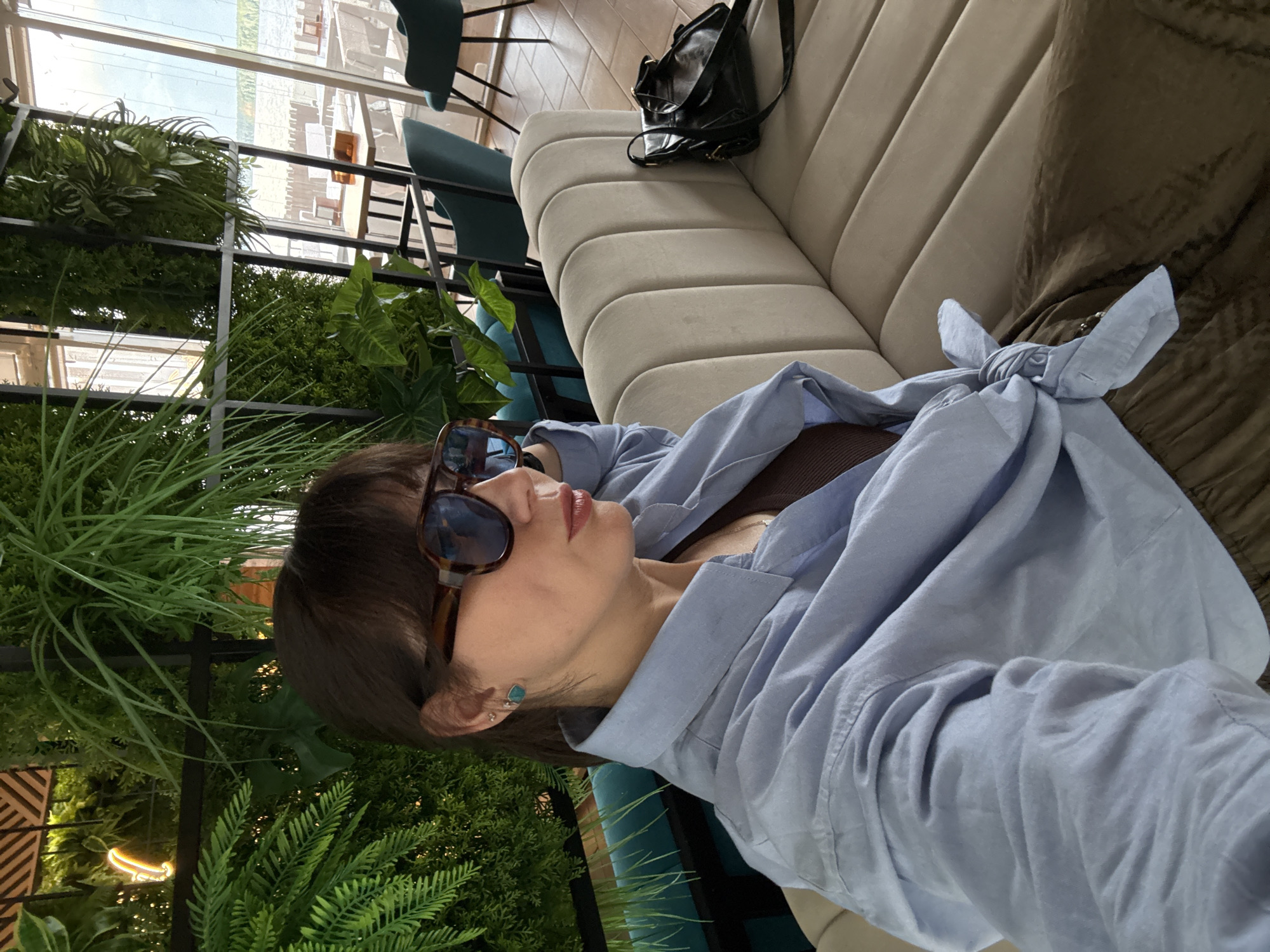A woman with dark hair and sunglasses sits on a couch in a restaurant. She is wearing a light blue button-down shirt tied at the waist and a brown top underneath. A black purse is on the couch next to her. The restaurant has a modern design with plants and large windows. The location is the Chaikovsky City Park in Chaikovsky, Russia.