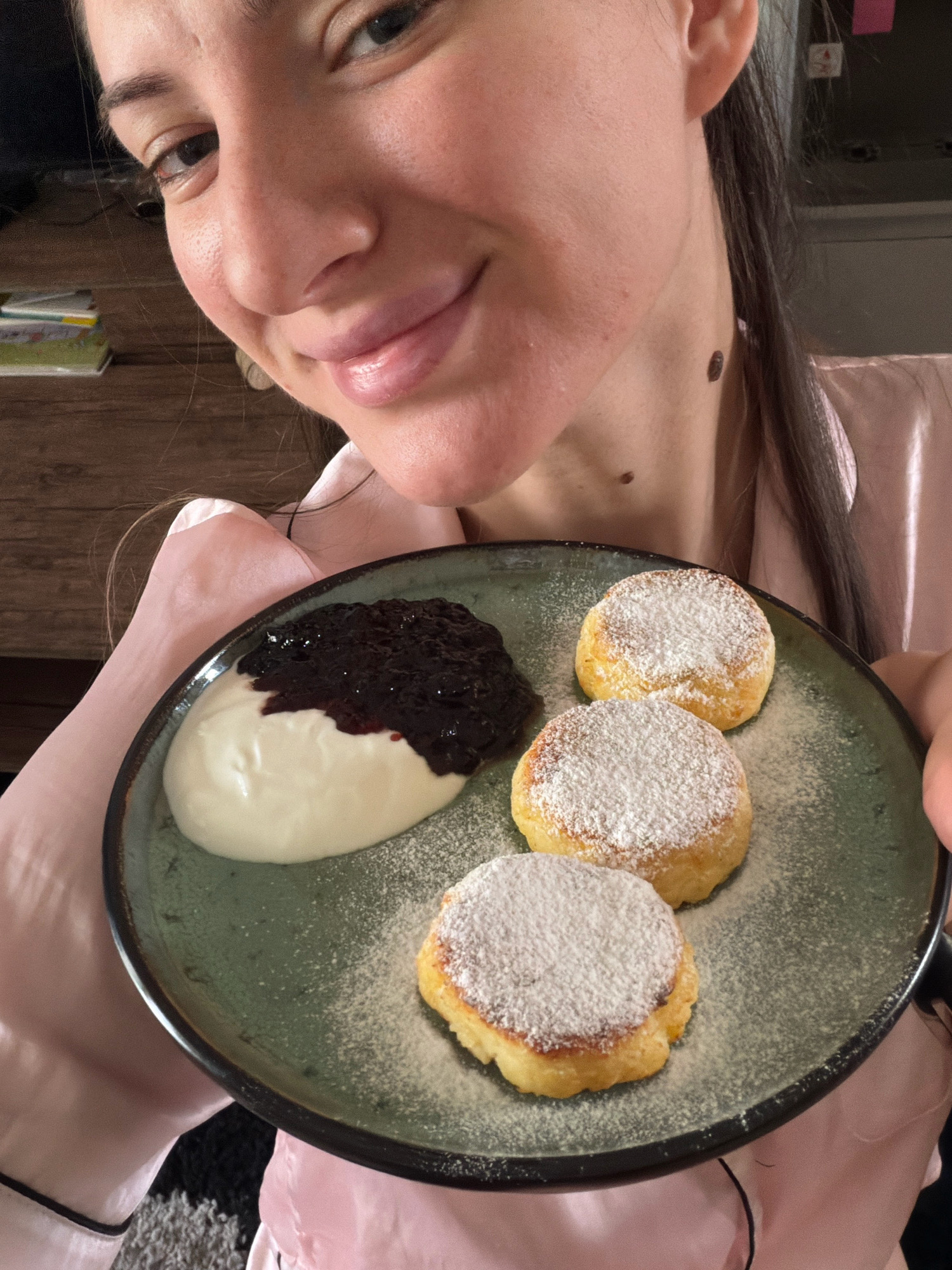 In a close-up indoor shot, a person wearing a pink silky garment holds a dark green plate containing a serving of syrniki, which are small cheese pancakes. The syrniki are dusted with powdered sugar. Beside the syrniki is a dollop of white cream topped with a dark jam, possibly blueberry or blackberry. The person's face is partially visible, smiling and looking towards the camera. The background suggests a domestic setting with wooden furniture and a glimpse of a television screen. There is no visible indication that this scene is located in Sharm El Sheikh, Egypt.