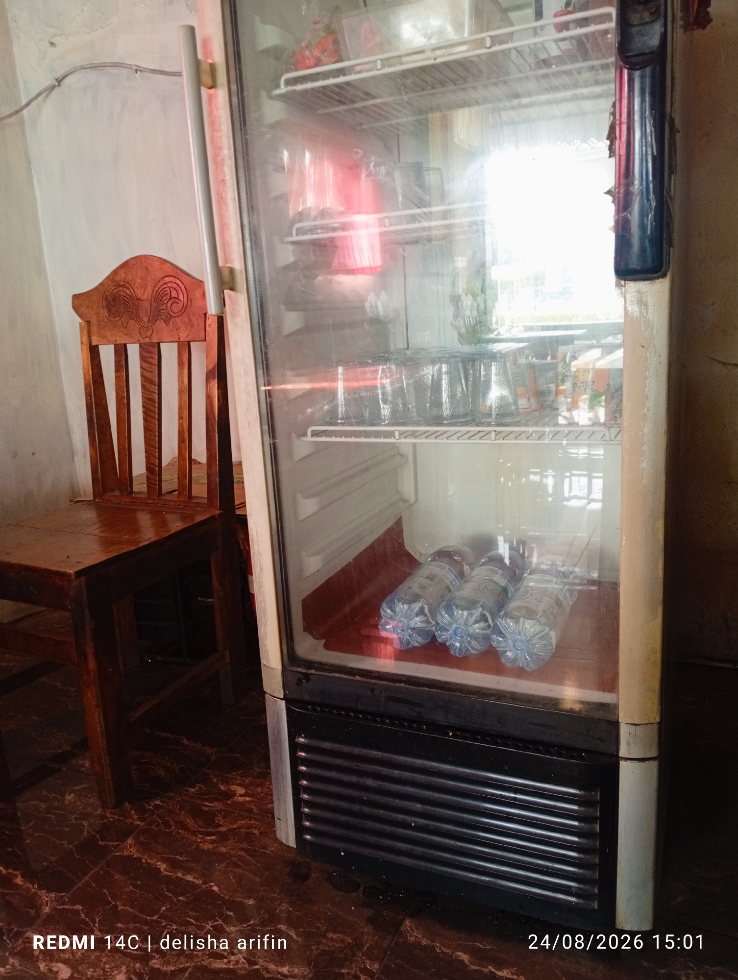 The image shows a tall, glass-doored refrigerator in what appears to be a storage area or small shop. The refrigerator is full of items, including bottles of water on the bottom shelf and various containers and possibly cans on the upper shelves. To the left of the refrigerator is a wooden chair with a carved backrest. The floor has a dark, marble-like pattern. The lighting in the image is bright, with a significant amount of light reflecting off the refrigerator door, obscuring some of the details inside. The background wall is a light color. Text overlays on the image indicate "REDMI 14C | delisha arifin" and "24/08/2026 15:01". The location is identified as Gambir Kuning, Indonesia.