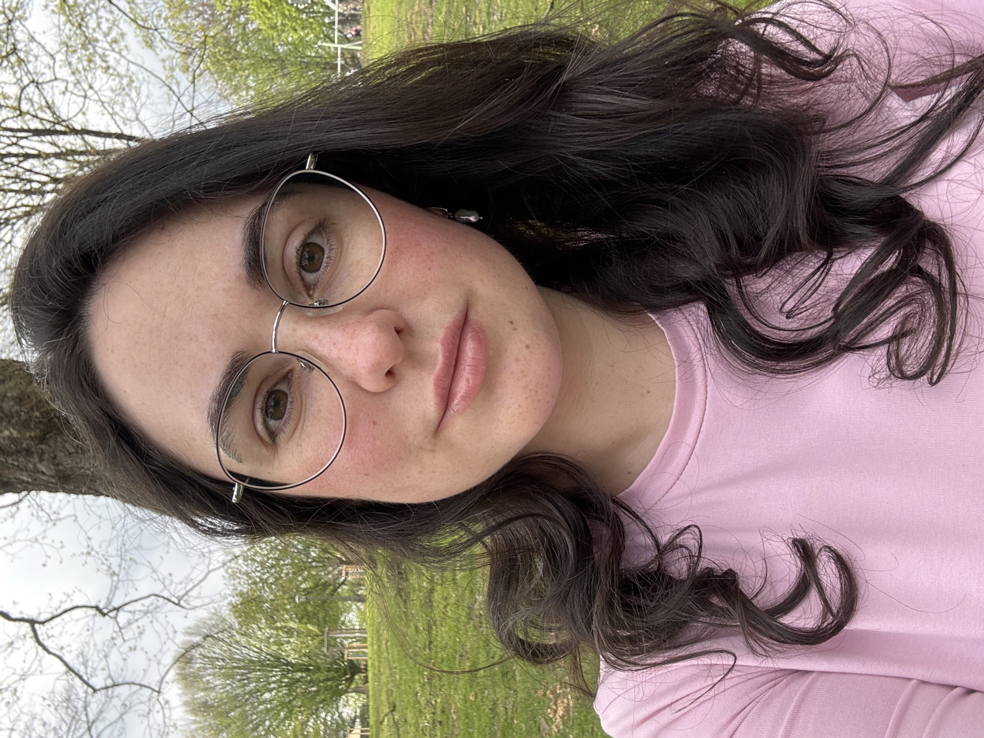 A young adult female is centered in a close-up outdoor portrait. She has fair skin with freckles, brown eyes, and dark curly hair. She wears round silver-rimmed glasses and a light pink t-shirt. The background shows a grassy field with trees in the distance. The image appears to be taken in a park or natural setting. The exact city and location within the city are unknown.
